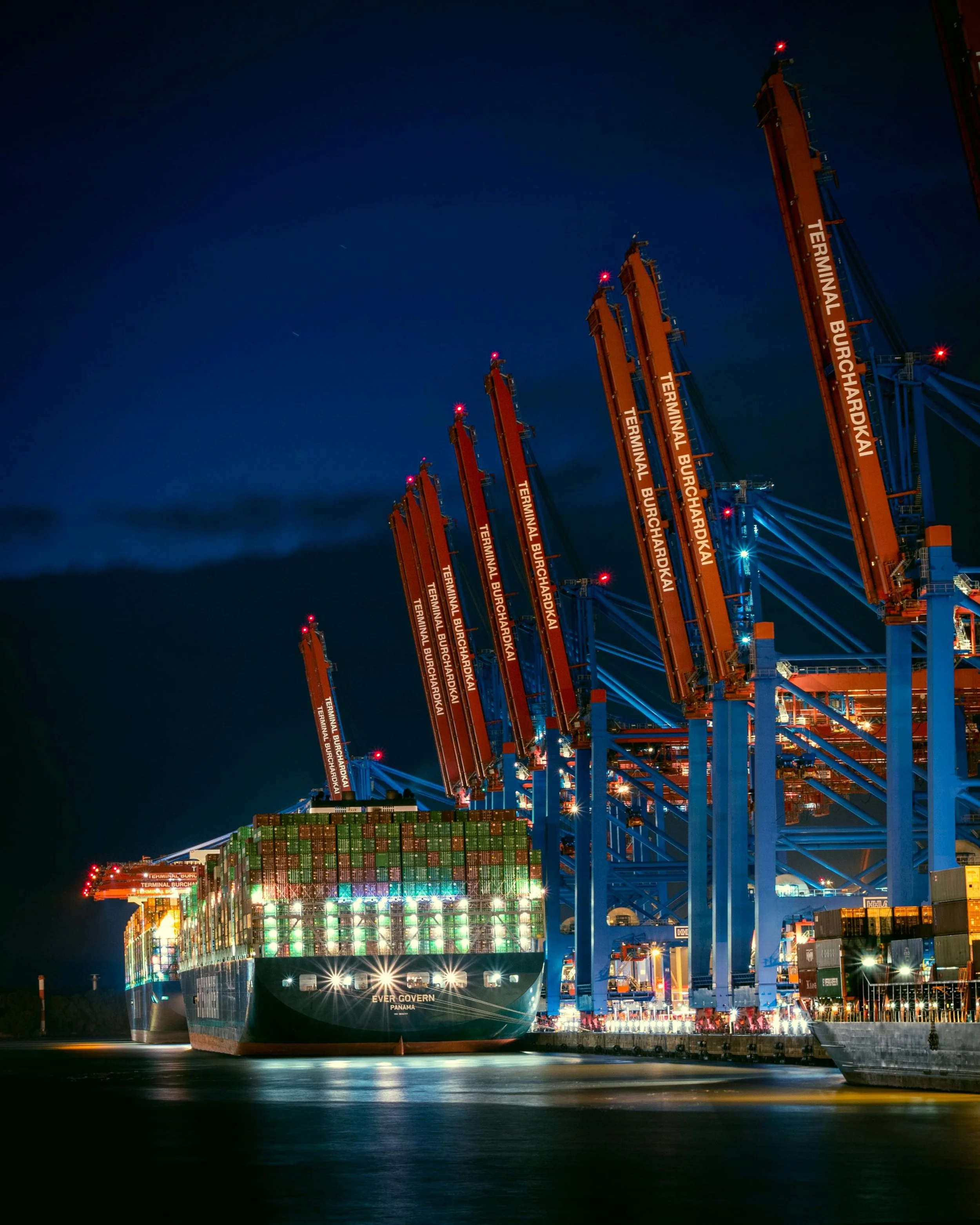 Large cargo ship named Ever Govern at Panama dock loaded with colorful shipping containers, docked at port with cranes labeled Terminal Burchardkai, illuminated by lights during nighttime.