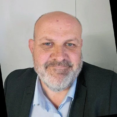 Portrait of a middle-aged man with a bald head and gray beard, wearing a dark blazer and light blue dress shirt, smiling at the camera.