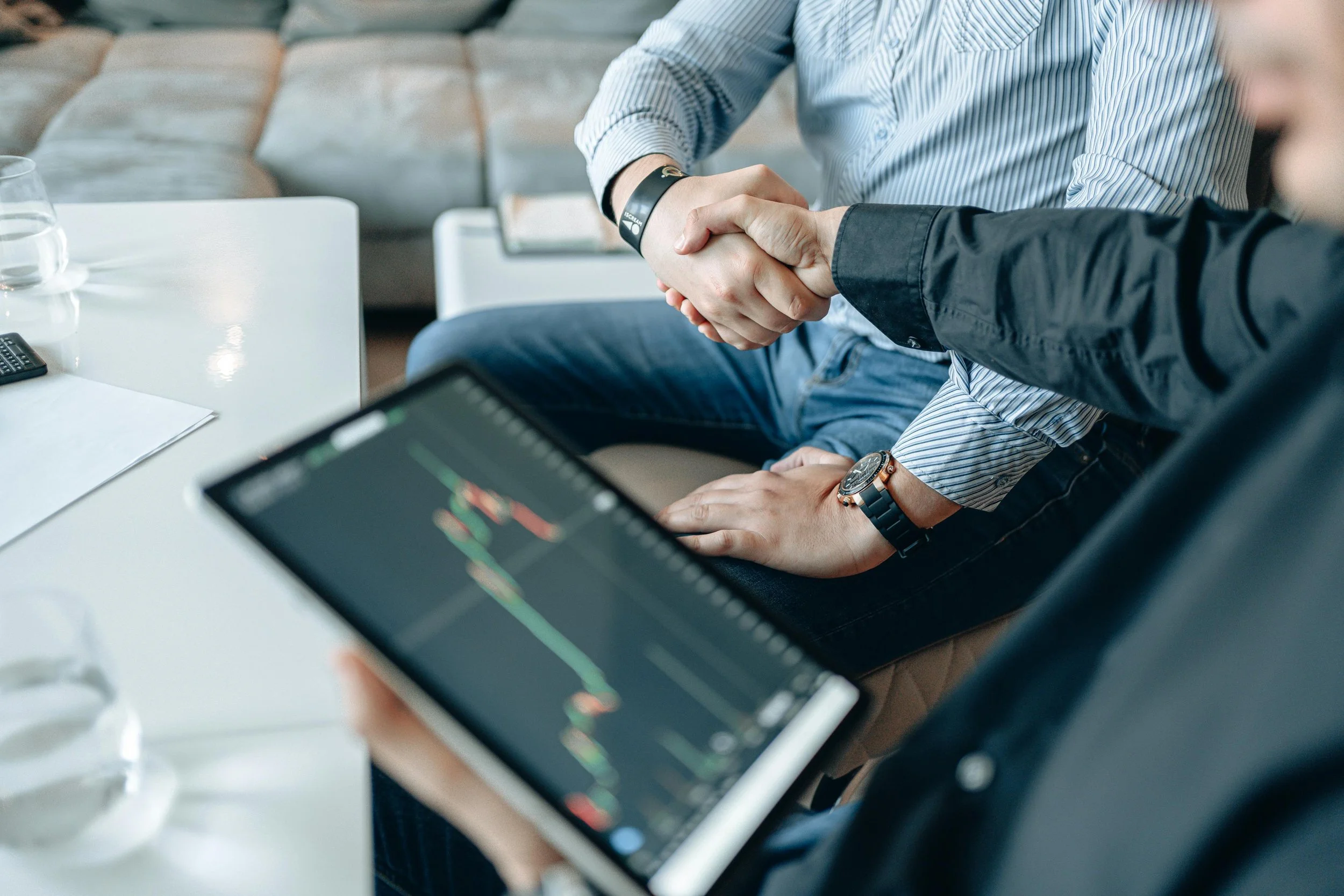 People shaking hands in a business meeting, with a tablet showing financial charts on the table.