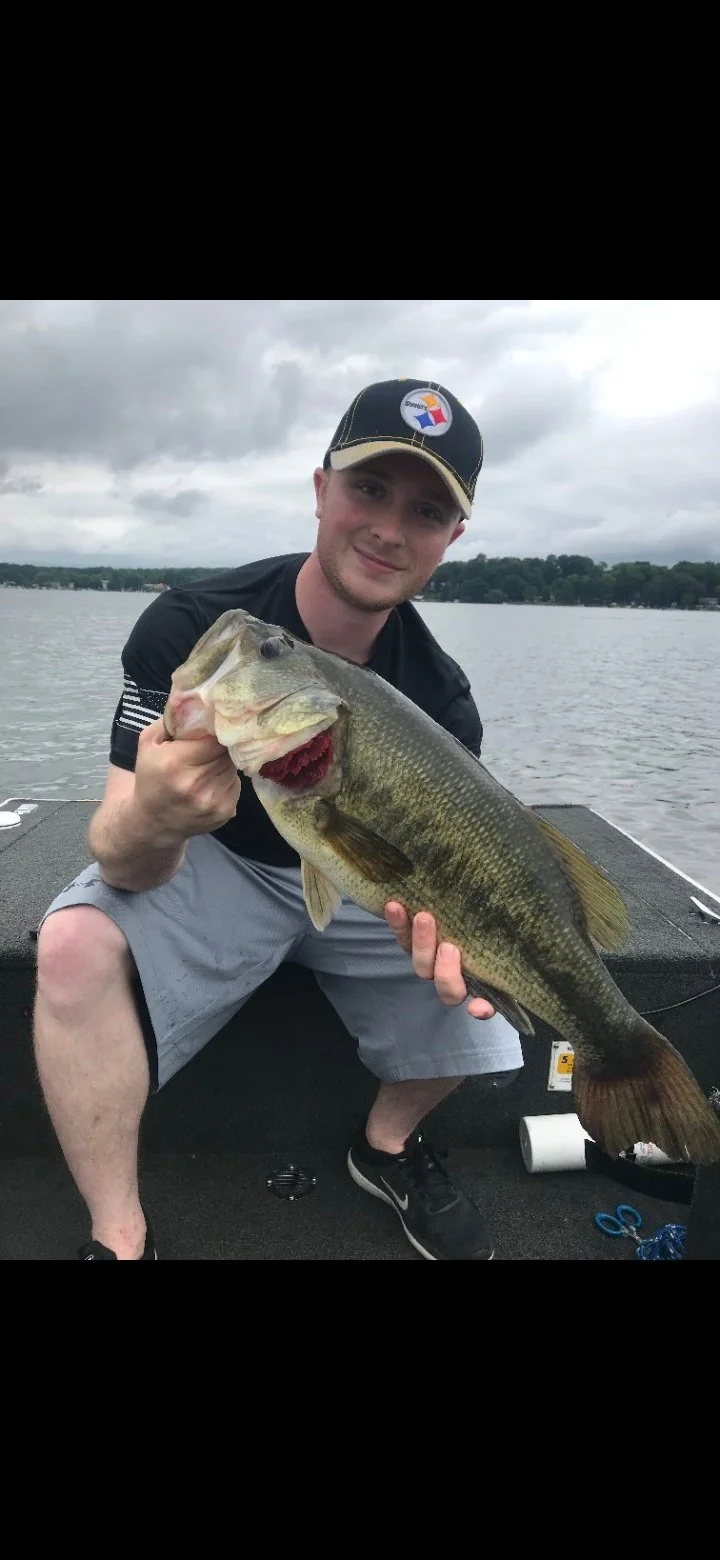 Young man holding a large bass fish on a boat, with water and cloudy sky in the background.