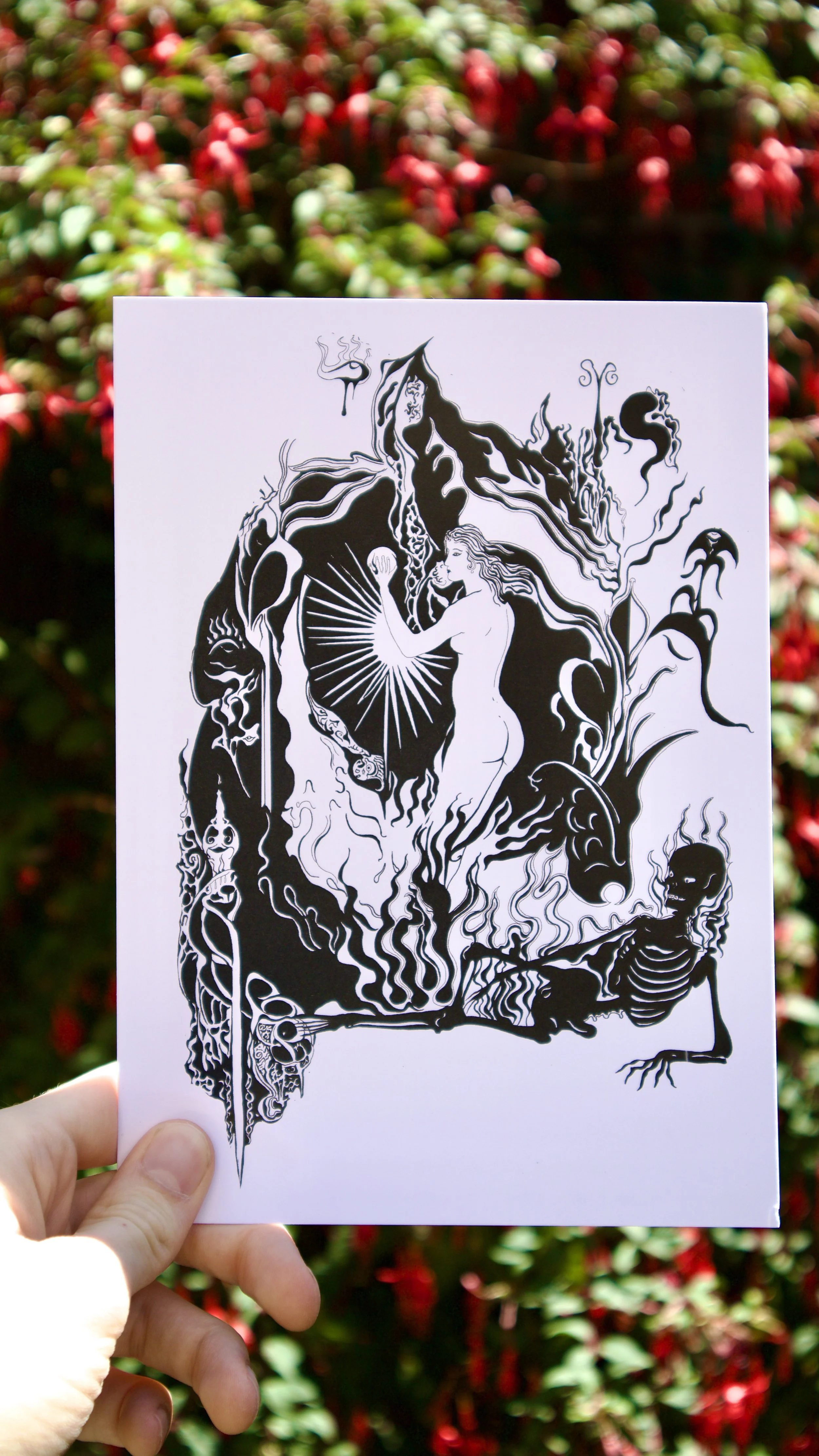 Eros and Thanatos print held against a floral background - a black and white gothic style pen and ink print featuring an art nouveau style woman and a flaming skeleton.