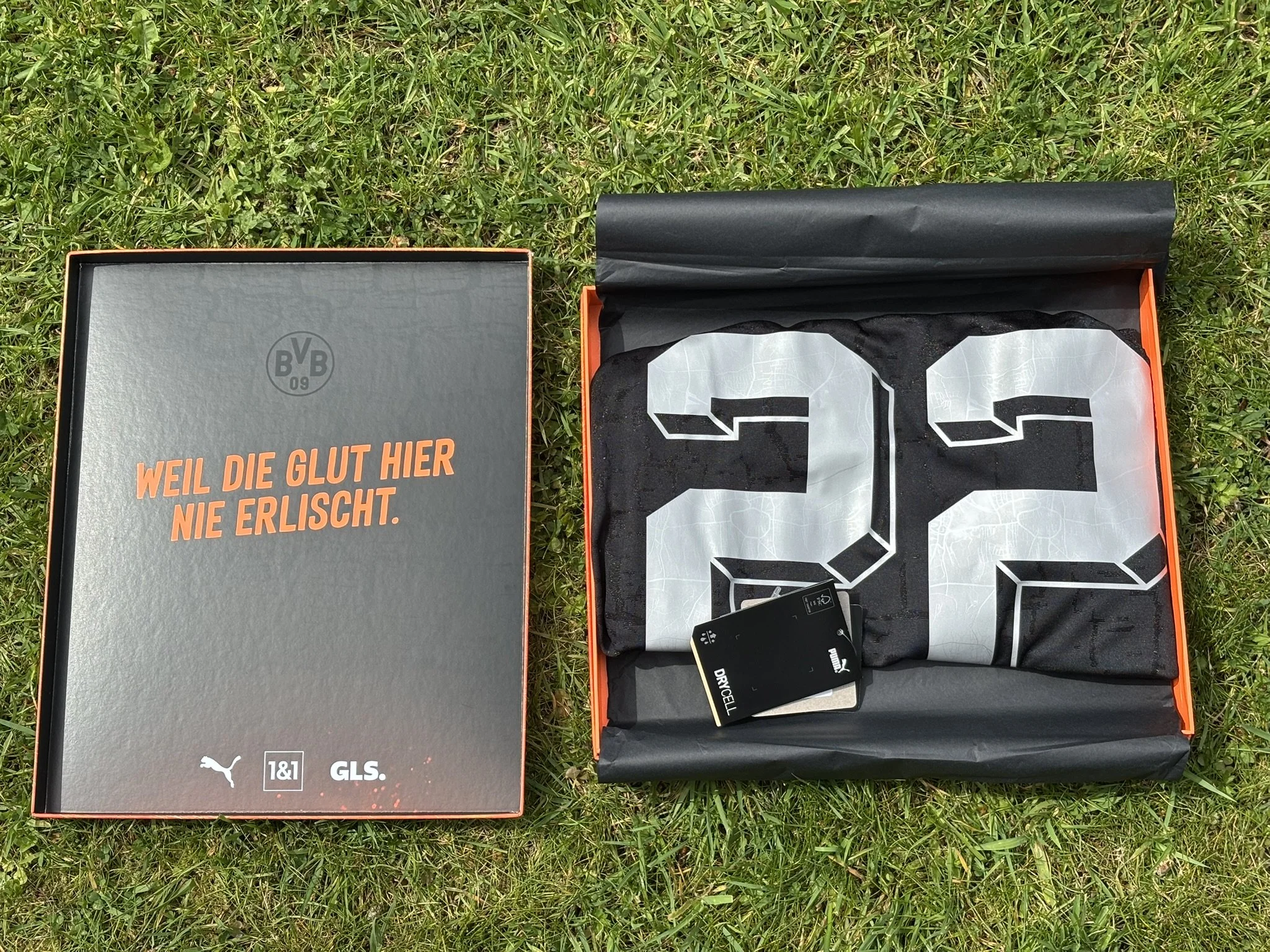 Open football shirt box with black and white clothing inside and paper lining, placed on grass, with German warning label and brand logos.