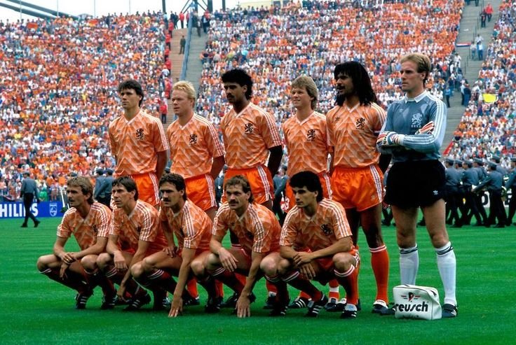 Dutch soccer team lined up for a pre-match photo on a green field. The team is wearing orange jerseys with white and orange stripes, and orange shorts. The goalkeeper is wearing a light blue jersey with darker blue stripes and dark shorts. There are large stands filled with spectators in the background.