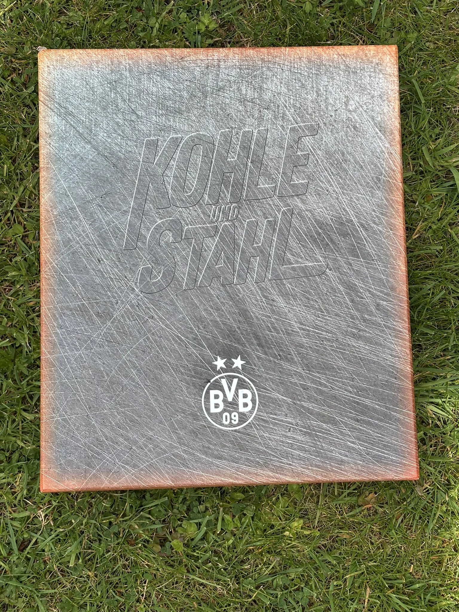 A rectangular metal object with a textured surface, displaying the text 'HOLE IN THE WALL' engraved on it, lying on green grass.