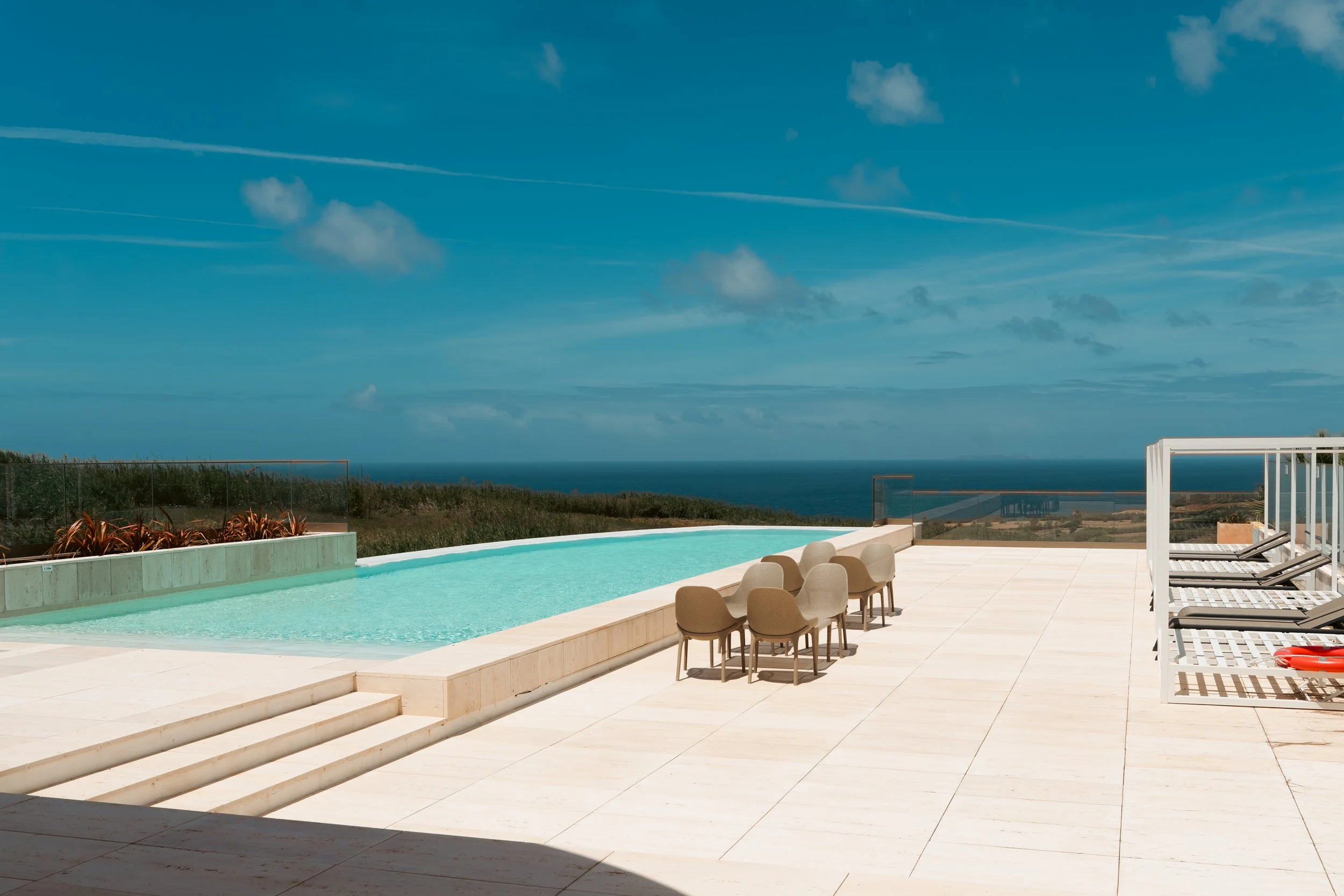A modern outdoor poolside area with lounge chairs, a pool, and a view of the ocean under a blue sky with clouds, East23 pool