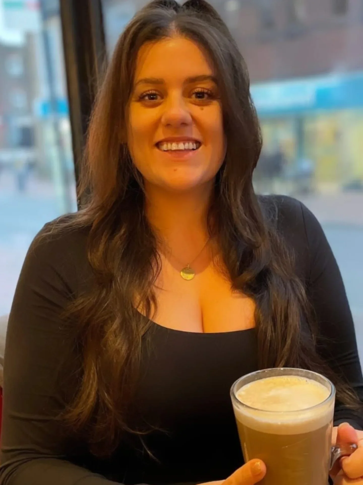 A young woman with long, wavy dark hair smiling and holding a mug of coffee in a restaurant or cafe setting.
