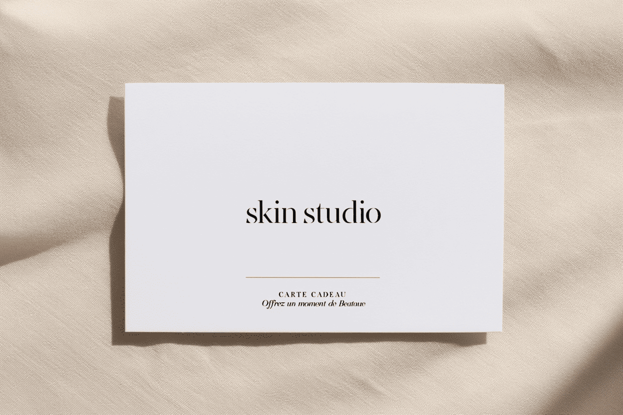 A white gift card with black text that reads 'skin studio' and smaller text that says 'CARTE CADEAU Offrez un moment de Beauté' placed on a beige textured fabric background.
