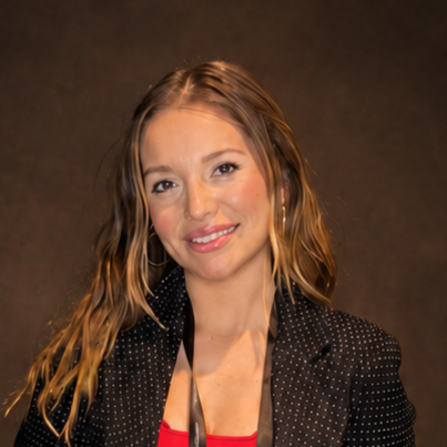 Portrait of a woman with wavy blonde hair, wearing a black polka dot blazer over a red top, smiling against a dark background.