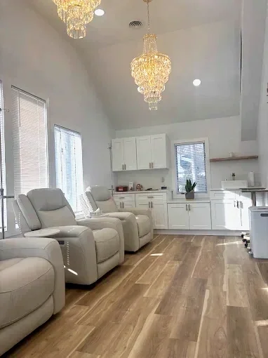 Living room with white leather recliners, white kitchen cabinets, and large windows, in a bright, modern space with hardwood floors and two chandeliers.