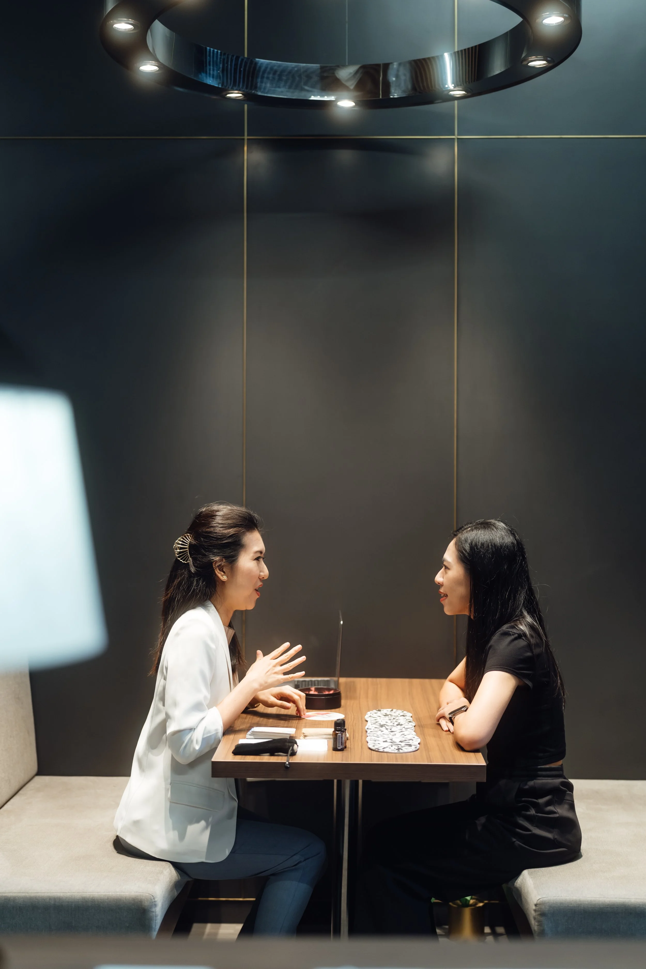 Two women are sitting across from each other at a wooden table, engaged in conversation. The woman on the left, wearing a white blazer and blue pants, appears to be speaking and gesturing with her hands. The woman on the right, dressed in black, is listening attentively. The setting is modern, with a dark wall, gold accents, and a circular ceiling light fixture above.