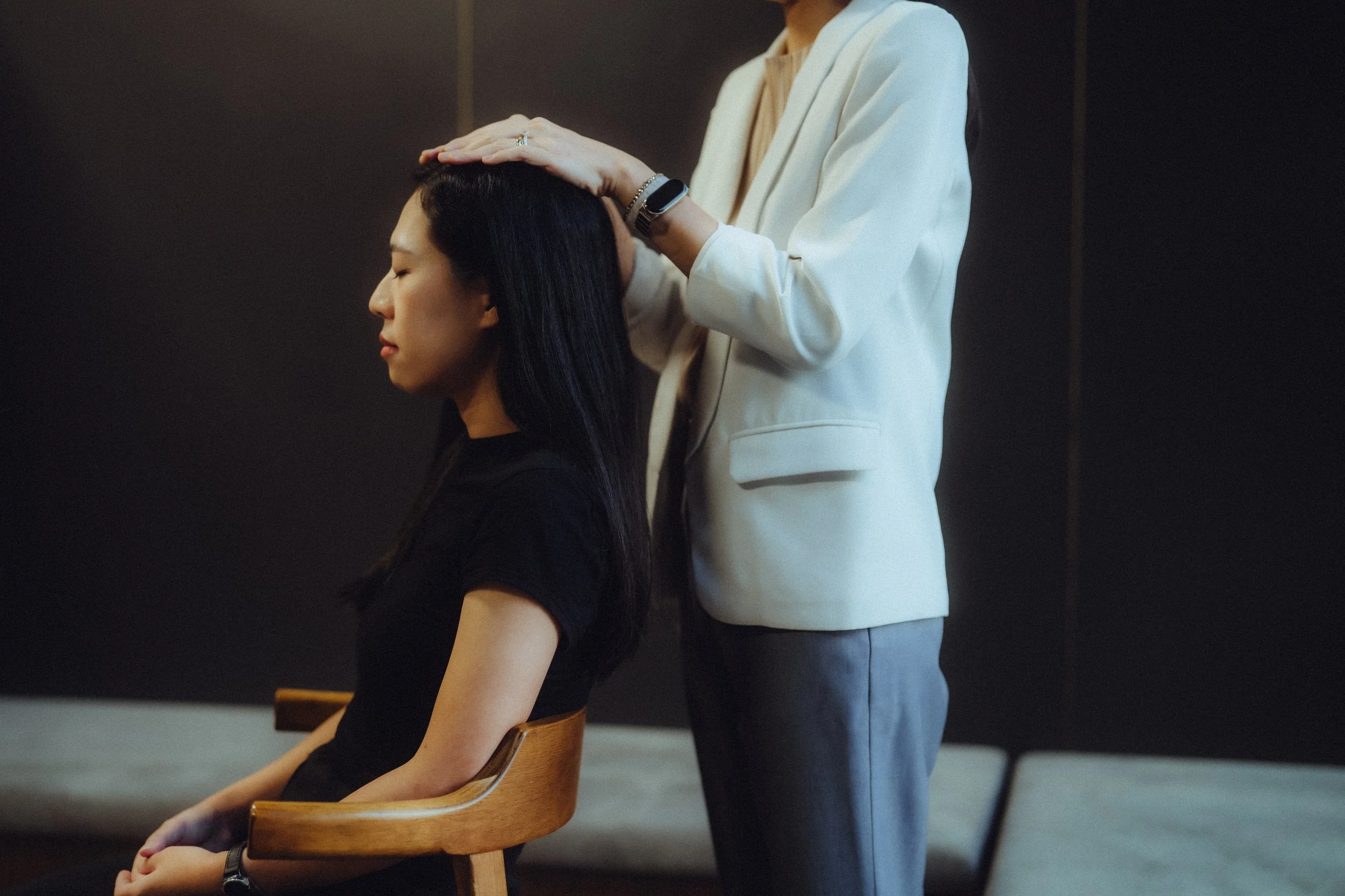 A woman with long dark hair sits with her eyes closed, while another person in a white blazer gently places their hand on her head during a massage or therapy session in a dimly lit room.