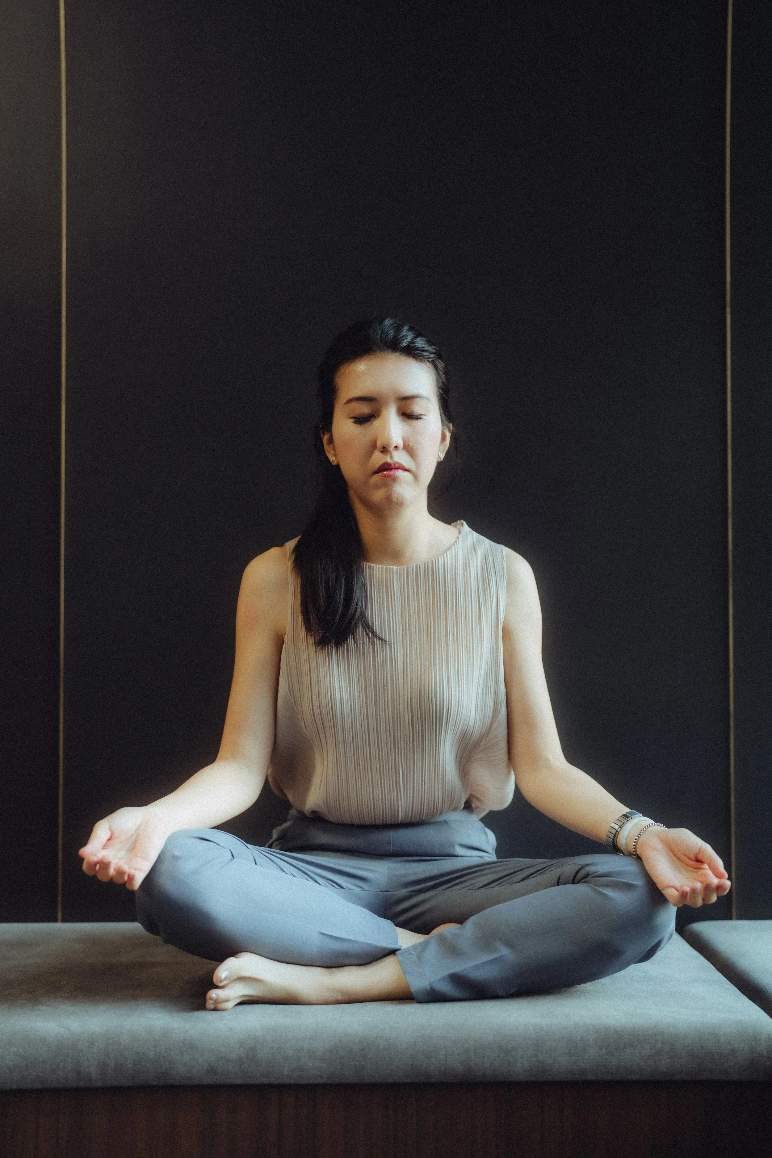A young woman sitting cross-legged on a yoga mat in a meditative pose with closed eyes, wearing a sleeveless top and gray pants, indoors against a dark wall with gold trim.