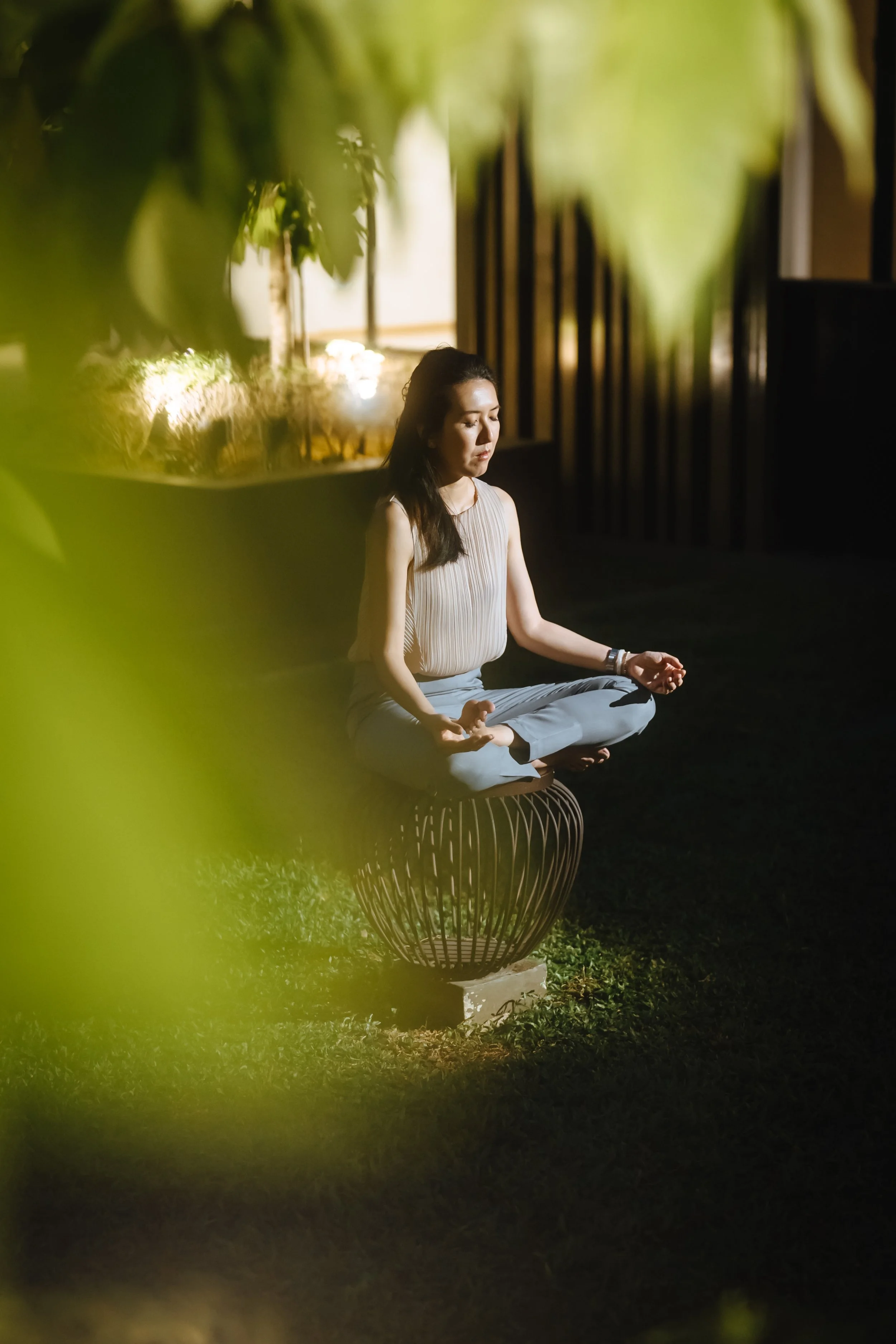 A woman practicing meditation outdoors at night, sitting cross-legged on a round woven stool with closed eyes, surrounded by greenery and illuminated by soft lighting.