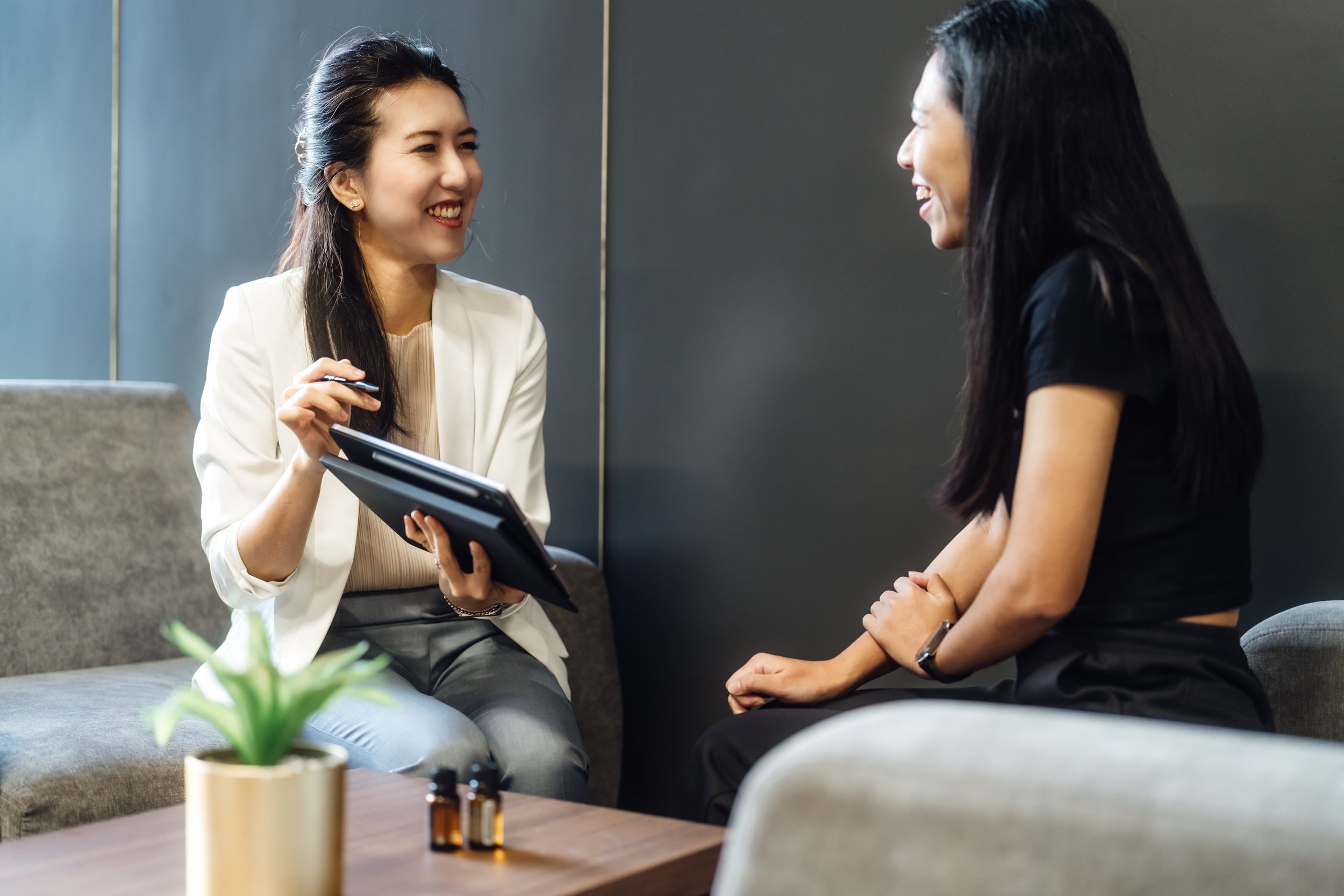 Two women are sitting and talking in a modern indoor setting. One woman, with dark hair and wearing a white blazer, is smiling and holding a notebook and pen. The other woman, with long dark hair and wearing a black shirt, is also smiling, and facing her. There is a small potted plant and a few bottles on a wooden table in the foreground.