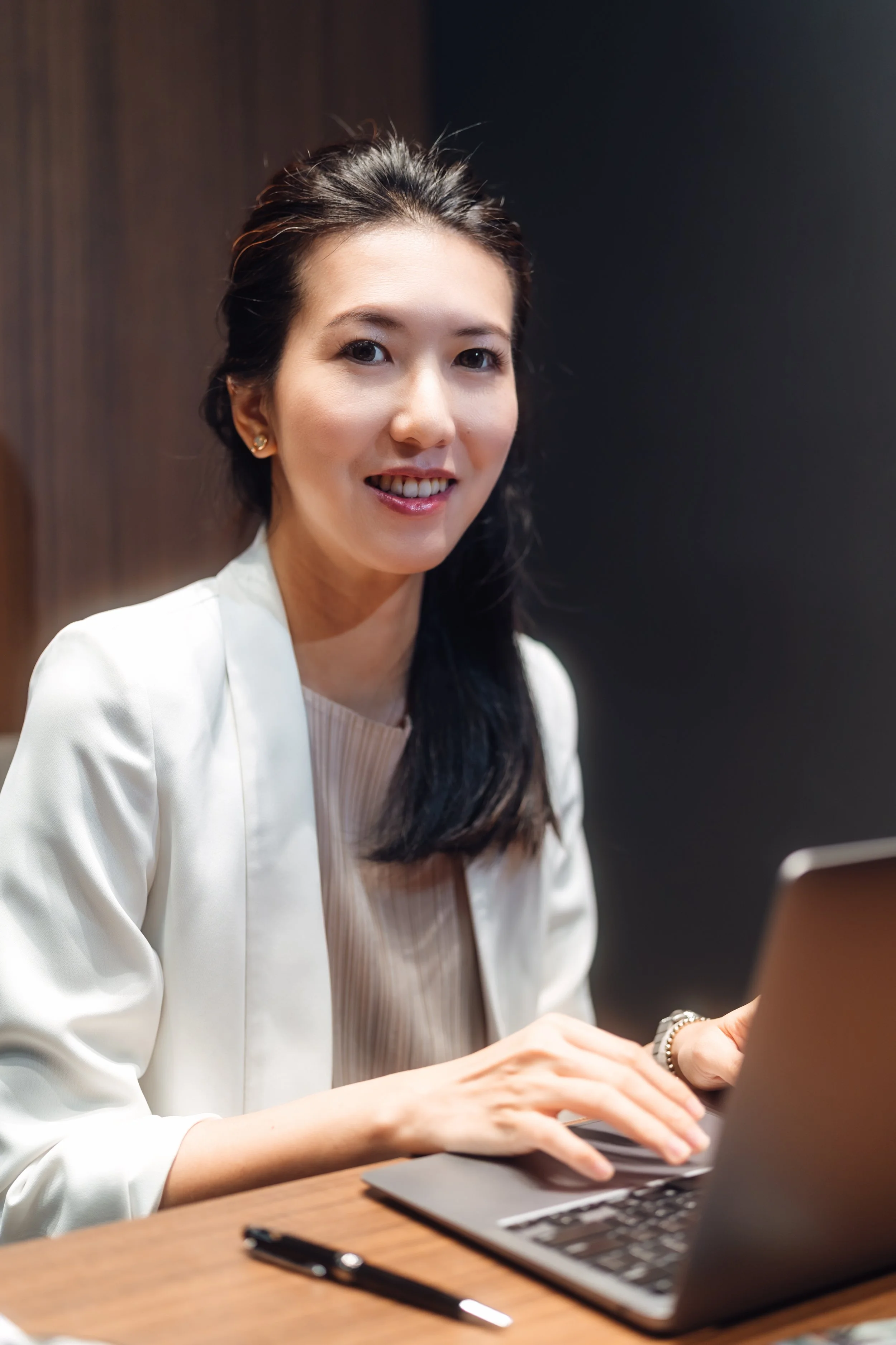 A woman with dark hair pulled back, wearing a light blazer and earrings, sitting at a wooden table using a laptop, with a pen and smartphone nearby.