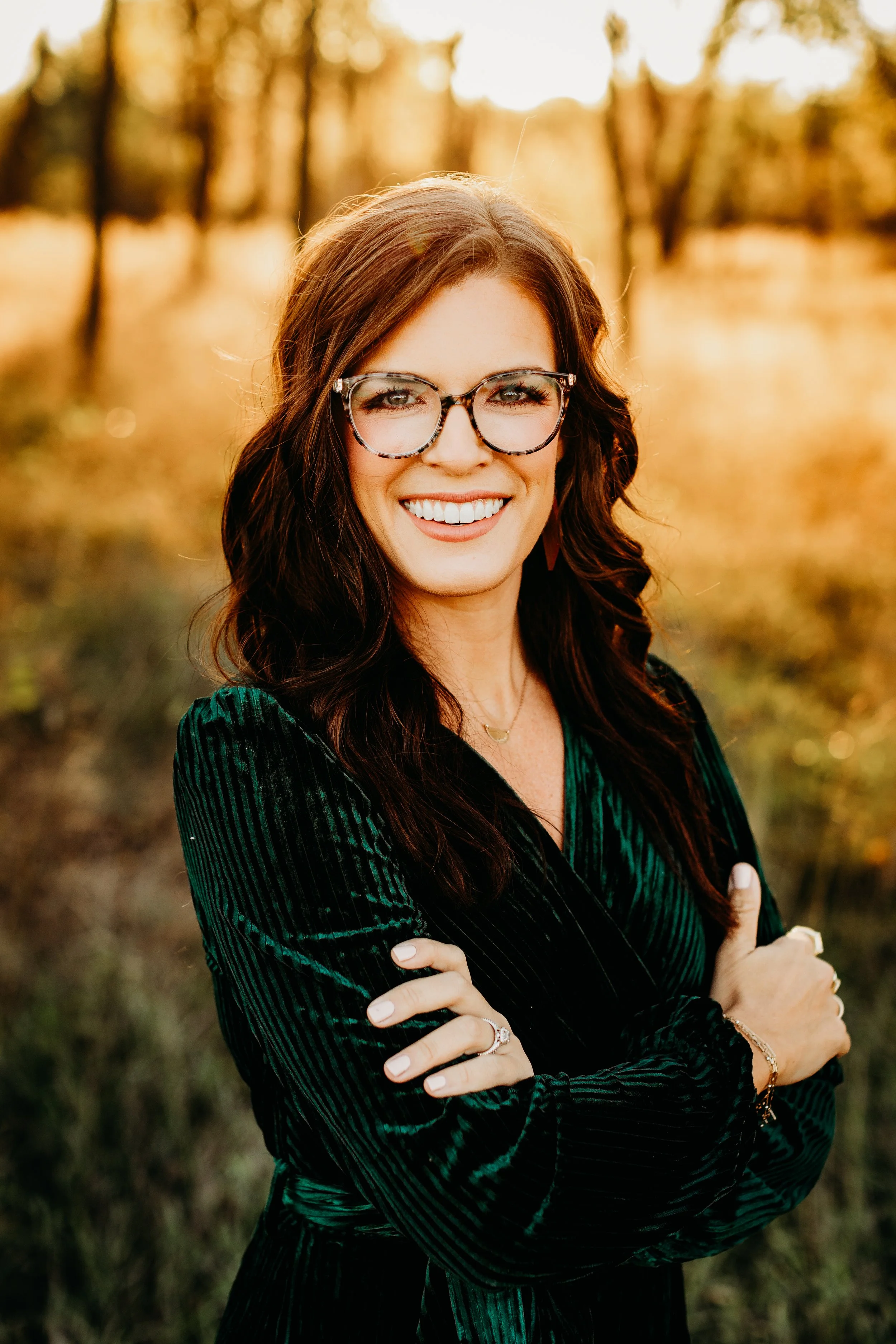 A woman with long brown curly hair wearing glasses with tortoiseshell frames, smiling outdoors during golden hour with trees in the background.