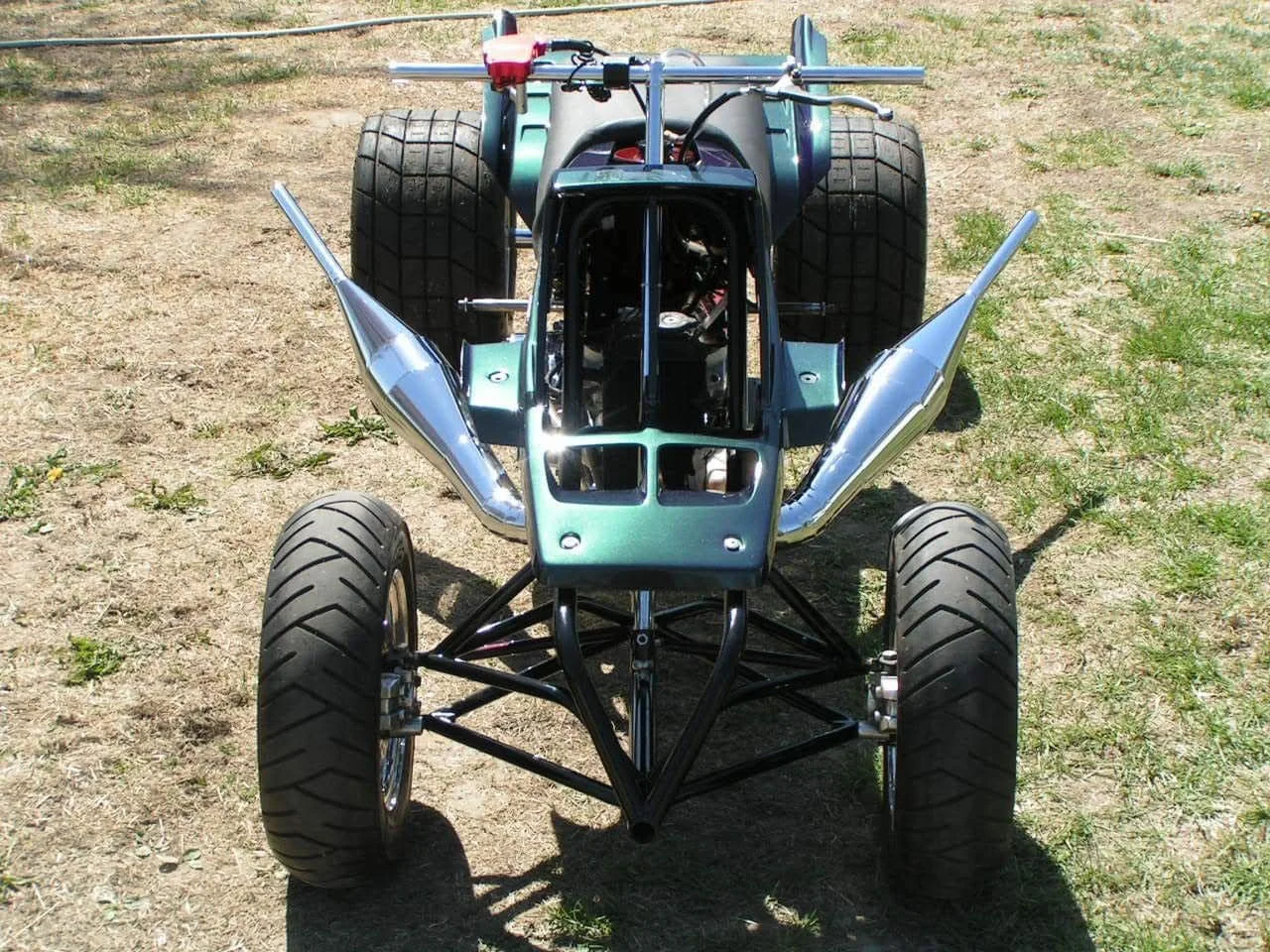 A front view of a vintage racing car with exposed wheels, aerodynamic fins on the sides, and a small cockpit, positioned on a grassy dirt area.