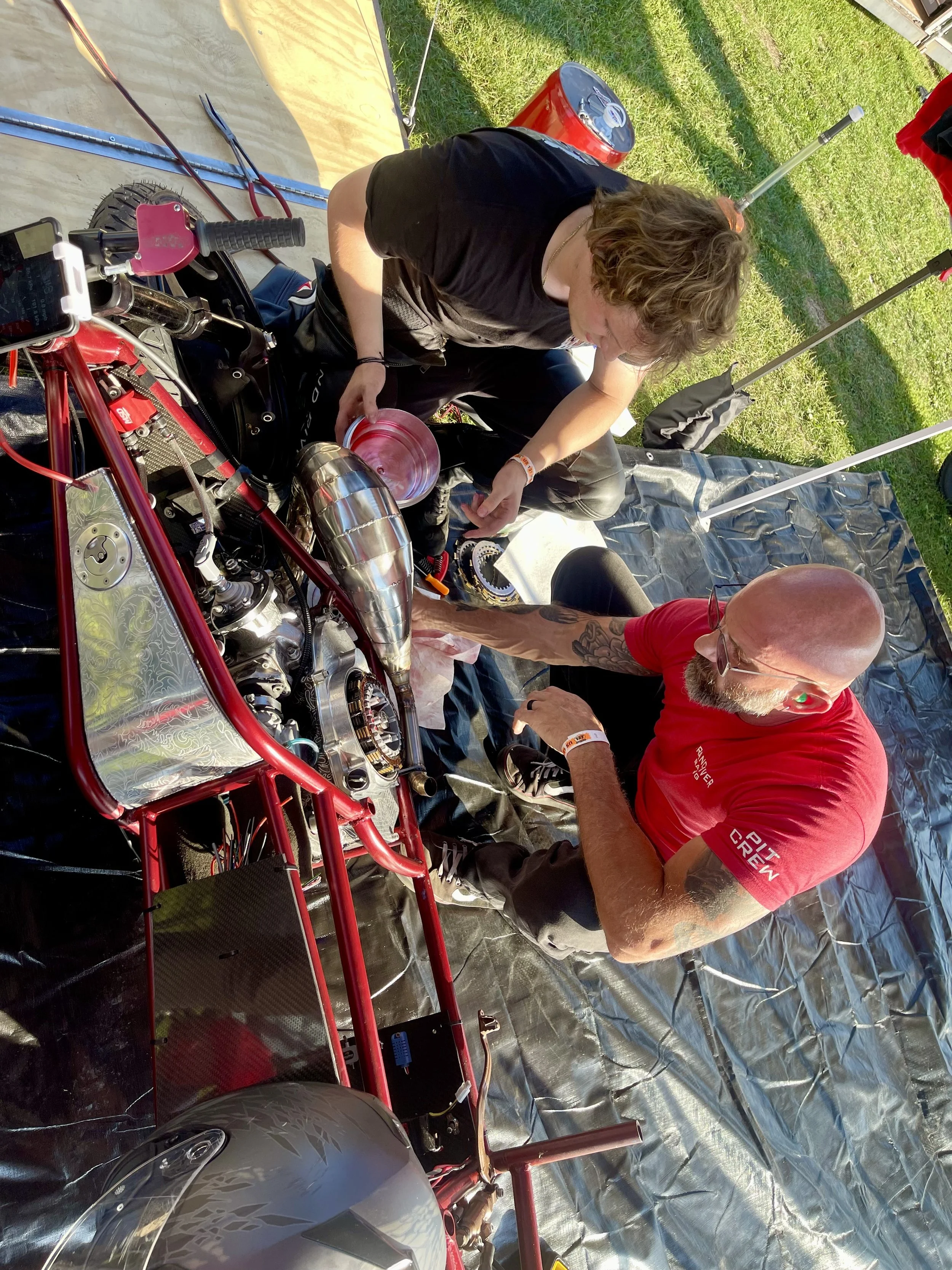 Two men working on a motorcycle engine outdoors during daytime, with tools and equipment around them, and a grassy background.