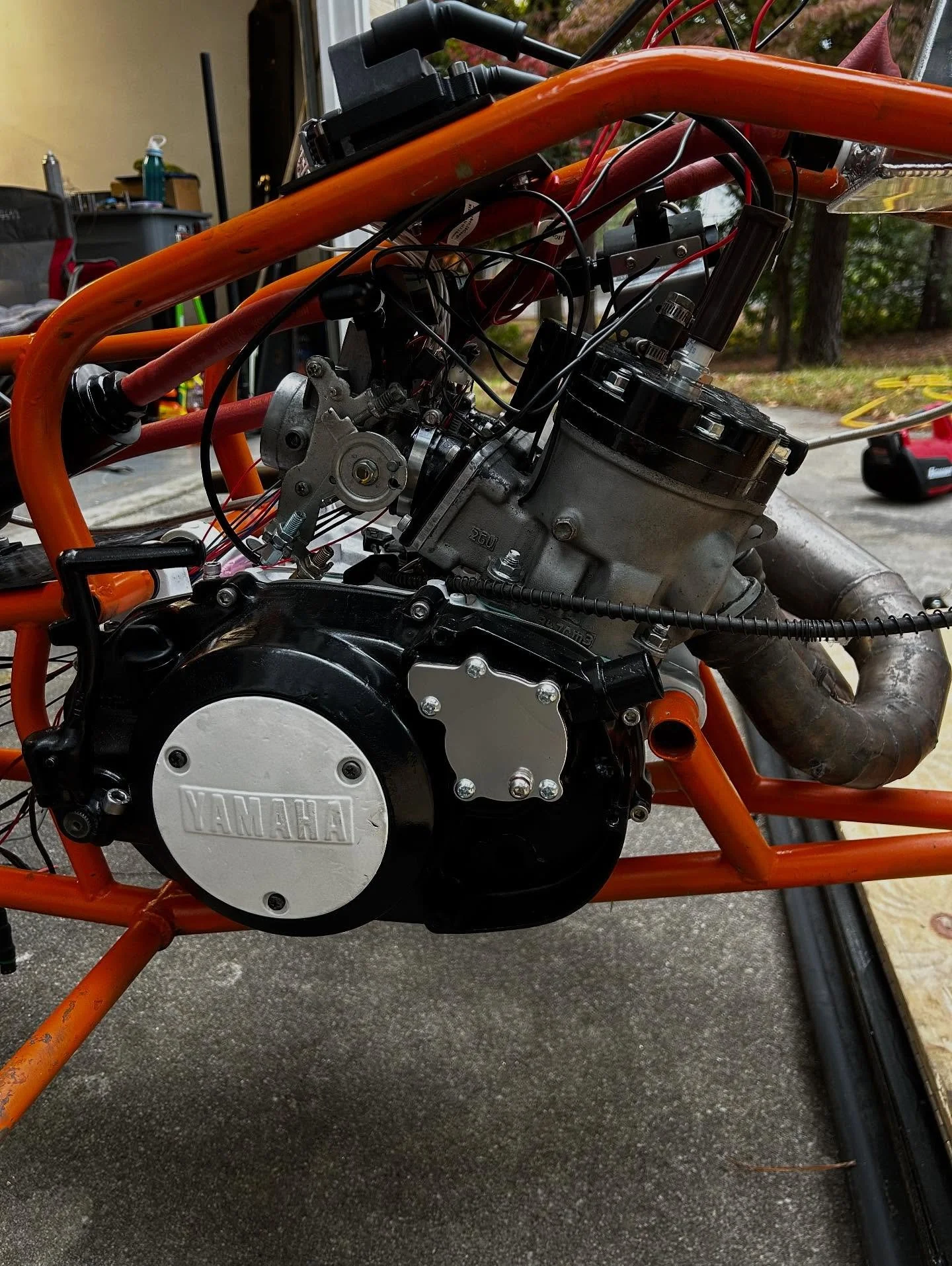 Close-up of a motorcycle engine with visible Yamaha logo, orange frame, visible wiring, and exhaust pipe, with a garage and trees outside in the background.