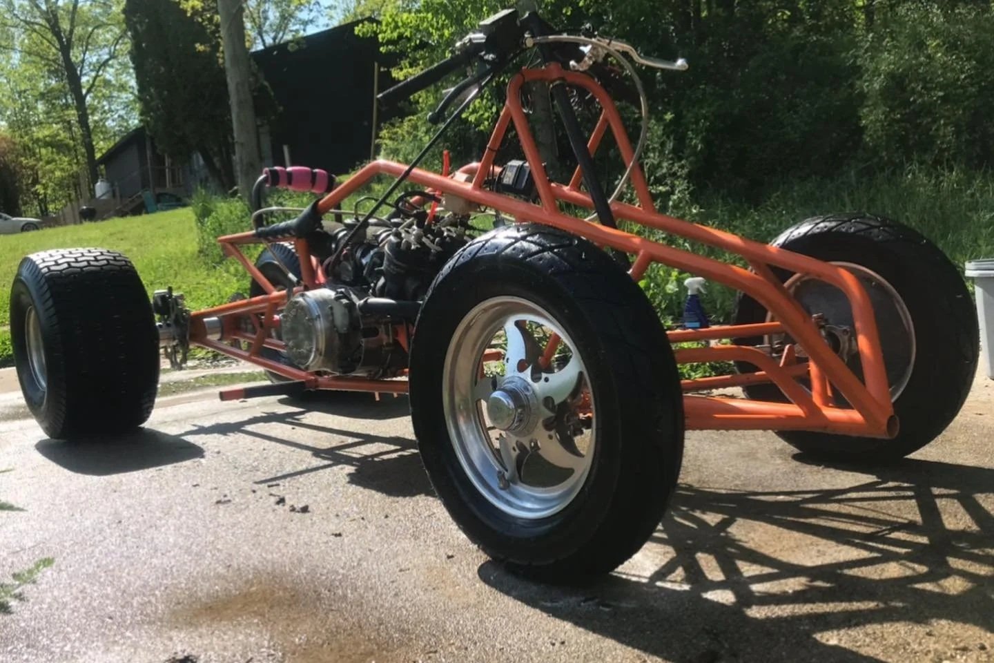 Front view of a partially assembled vintage-style race car chassis with boxed frame, large rear tires, smaller front tires, and visible engine components, on a paved surface outdoors.
