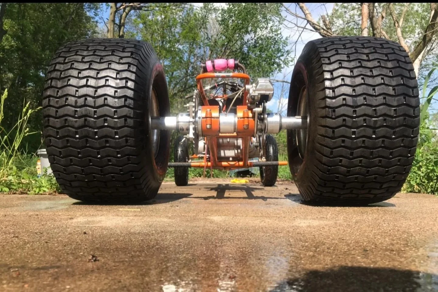 Close-up of a vintage race car's undercarriage with large tires, visible mechanical components, and a clear sky with trees in the background.