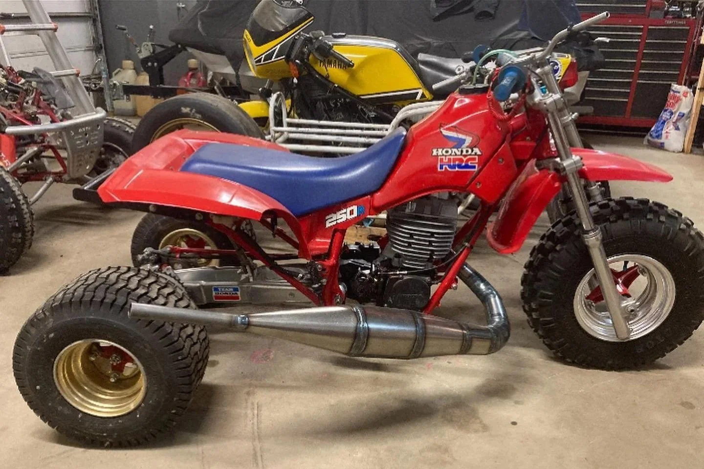 A vintage Honda motorcycle with off-road tires, red body, blue seat, and a large exhaust pipe, inside a garage with other motorcycles and tools.