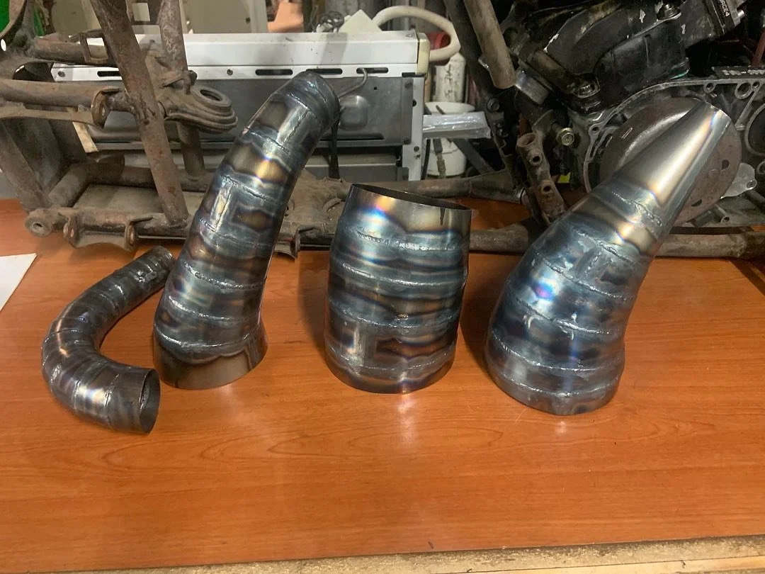 Four metal exhaust pipes with heat discoloration, displayed on a wooden surface with mechanical parts and tools in the background.