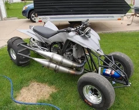 A custom-built, modified quad bike with a motorcycle engine, large rear tires, and a small front frame, parked on grass near a trailer.