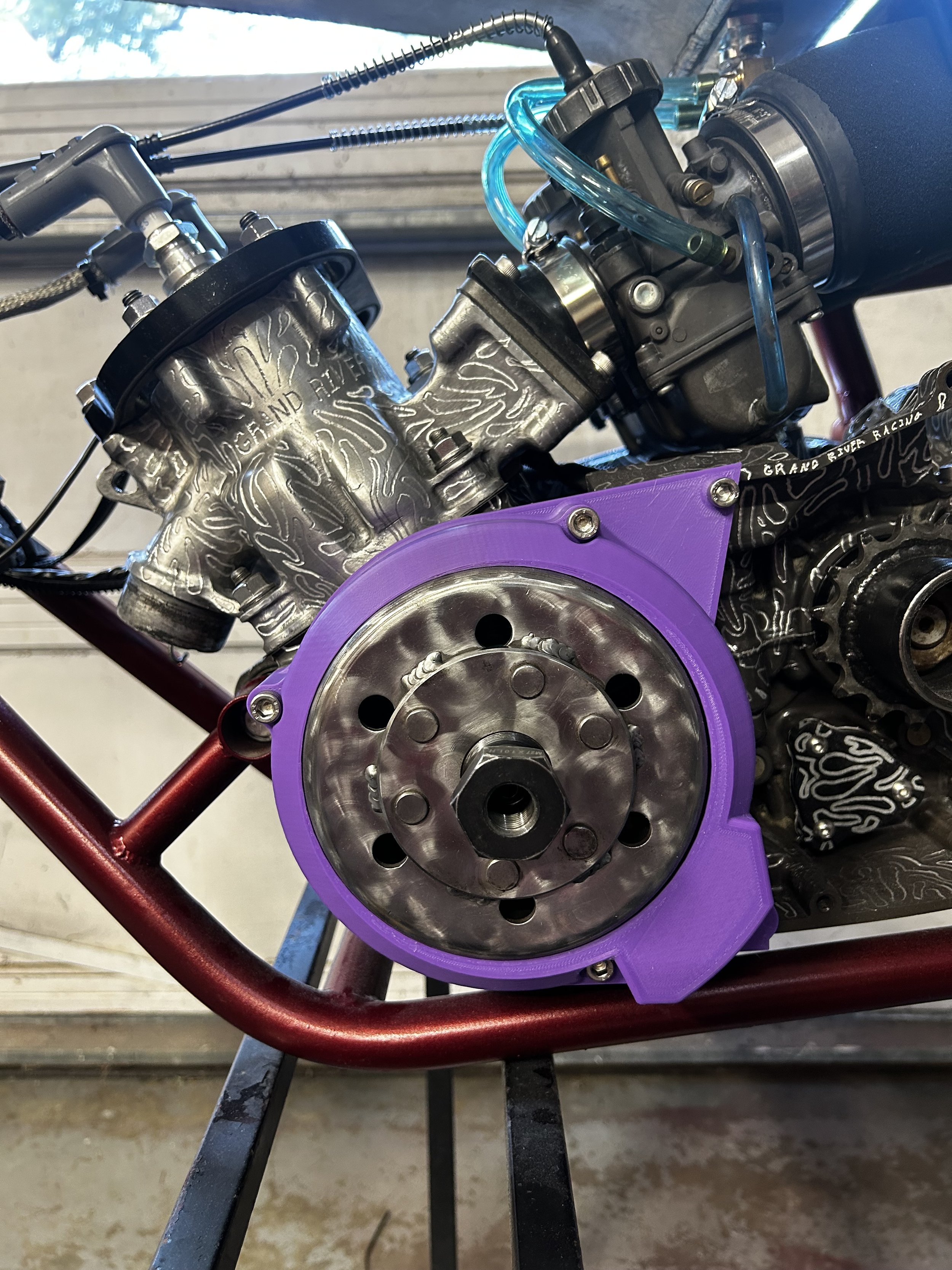Close-up of a race car engine with visible metal components, black hoses, and purple custom parts, mounted on a black frame with a red support bar.