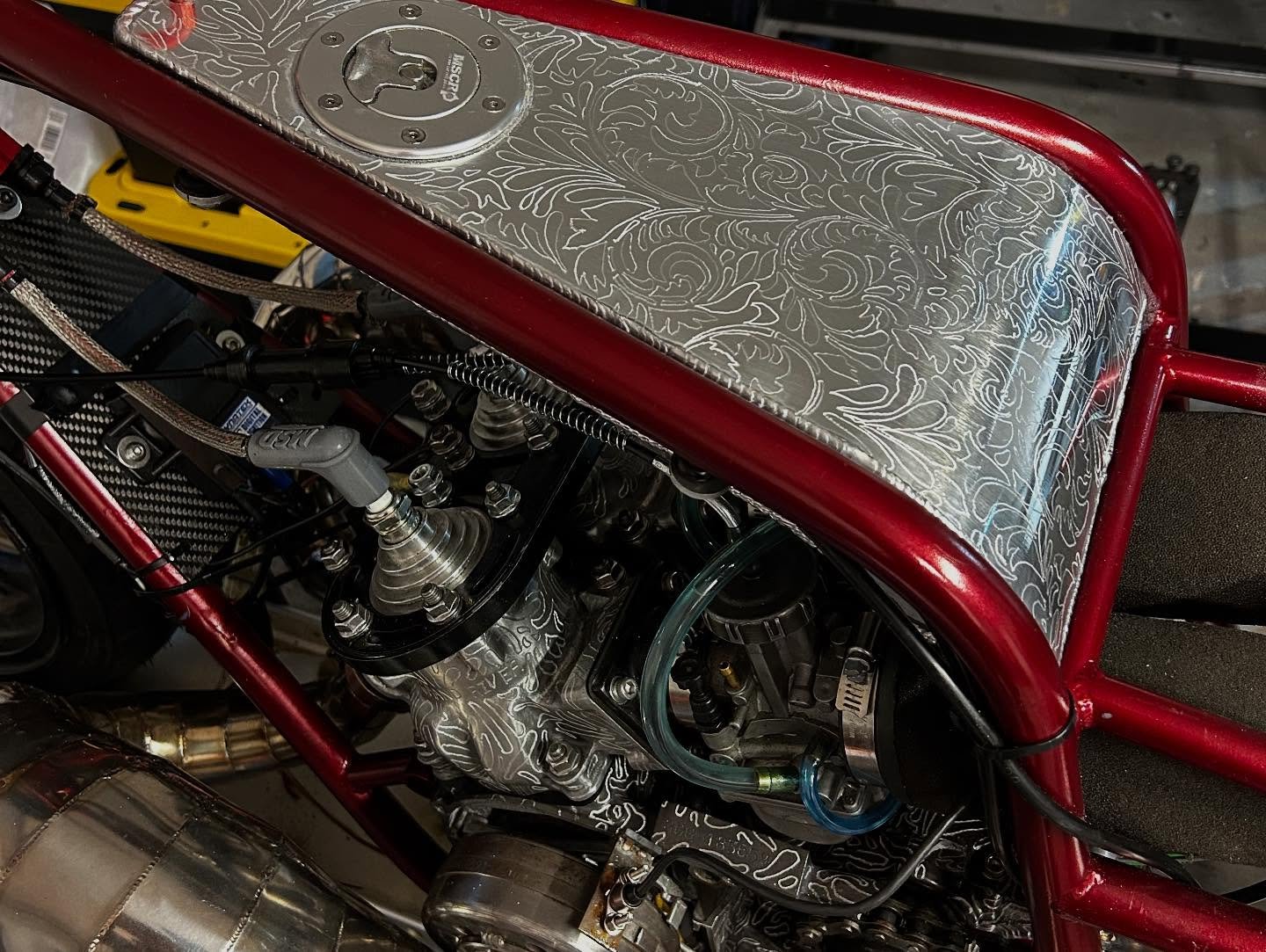 Close-up of racing car engine with elaborate engraved metal components, red frame, and carbon fiber parts.