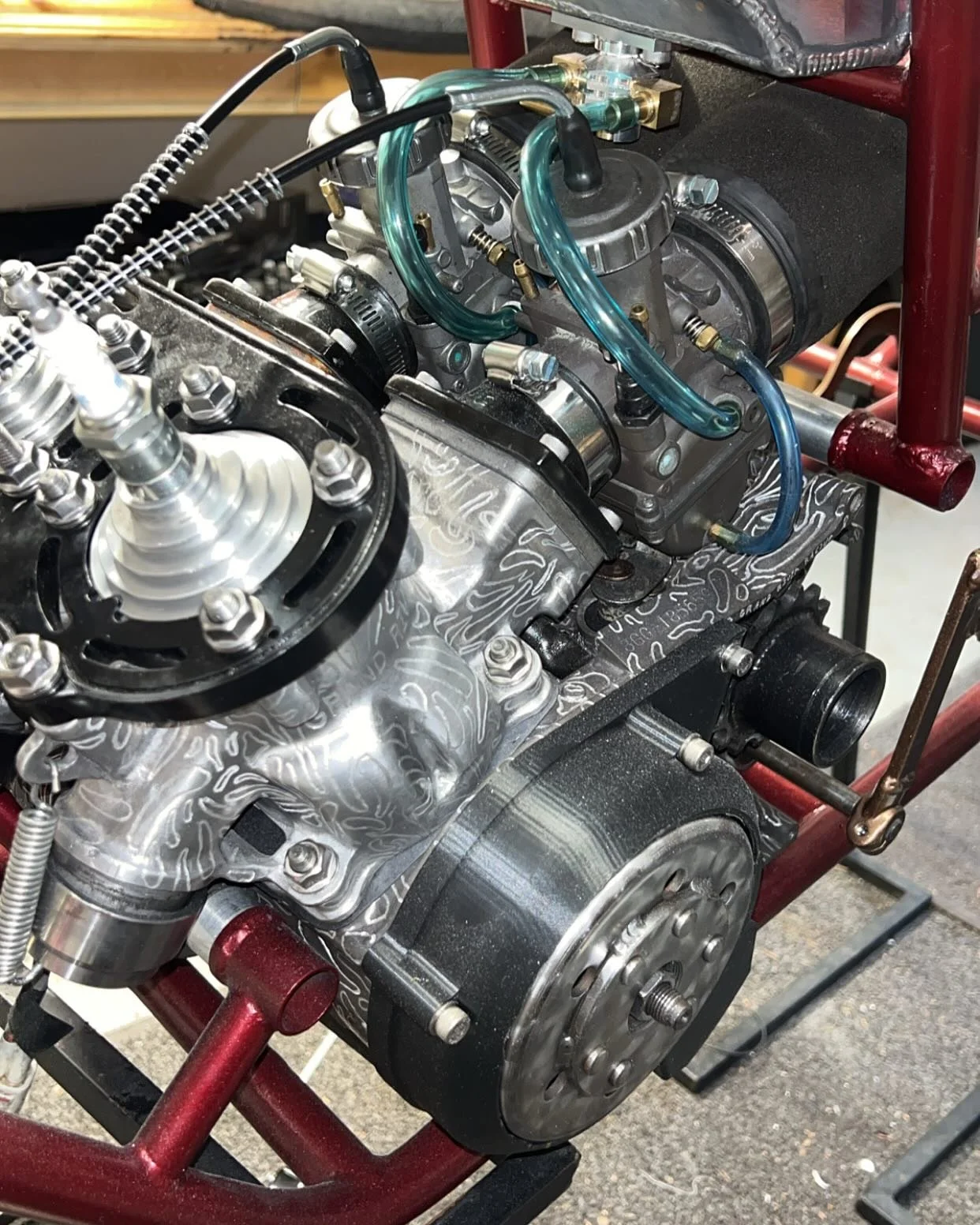 Close-up of a racing engine mounted on a red metal frame, with visible valves, hoses, and shiny metallic components.