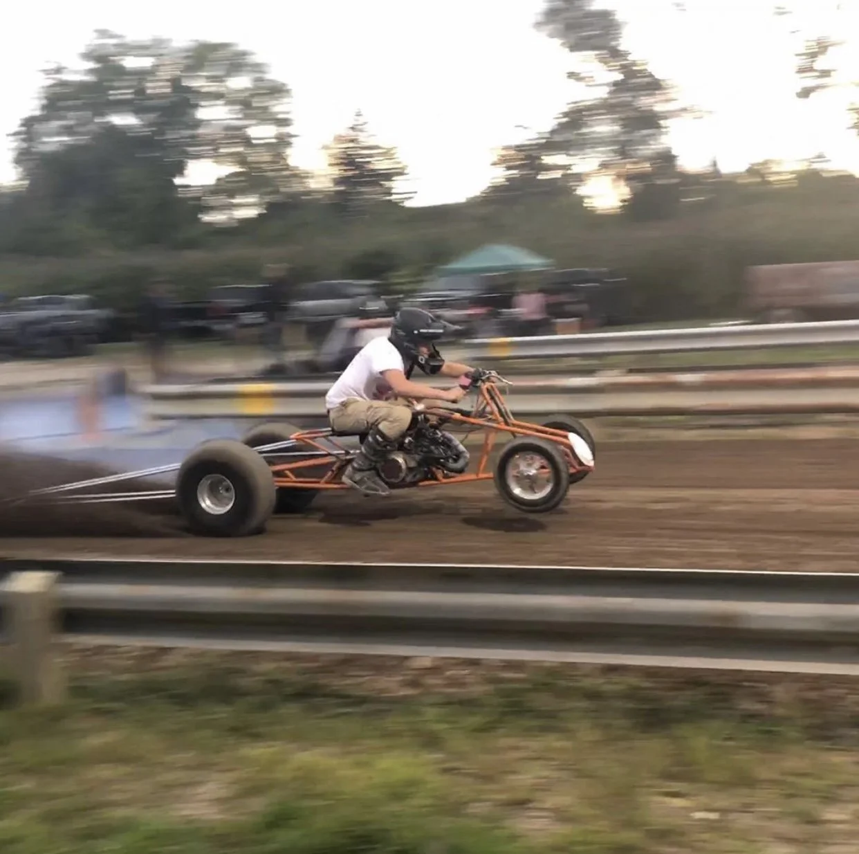 Person wearing a helmet and white T-shirt riding a drag racing motorcycle on a dirt track, with a background of trees and parked cars, motion-blurred due to speed.