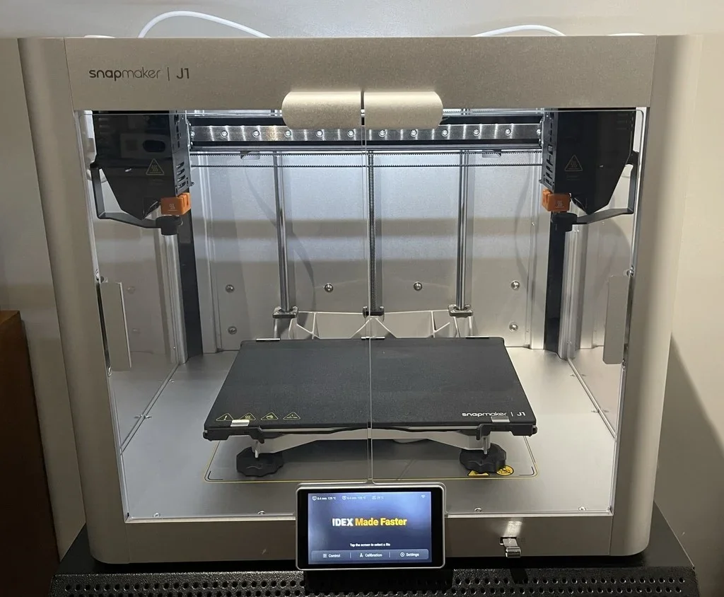 A Snapmaker J1 3D printer with a black print bed, placed inside a metal frame, with a small touchscreen interface displaying "IDEX Made Faster."
