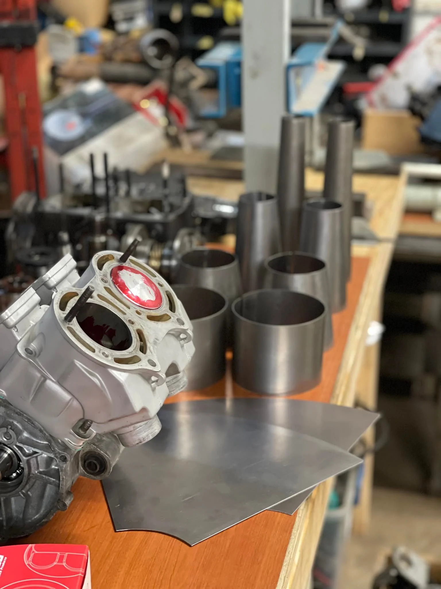 Close-up of a mechanical engine part, likely a cylinder head, on a workbench with metal cylinders and tools in the background.