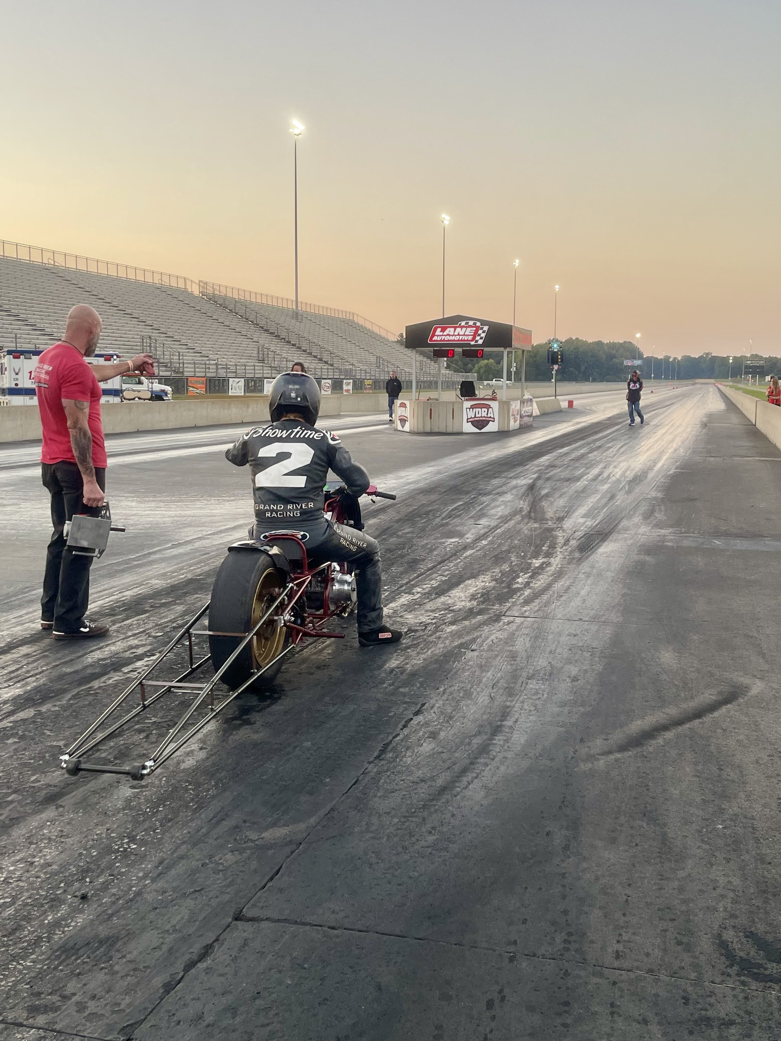 A drag racing track with a person on a racing bike, a man in a red shirt, and a woman in racing gear. The track is prepared for a race with a starting line, and the grandstands are empty.