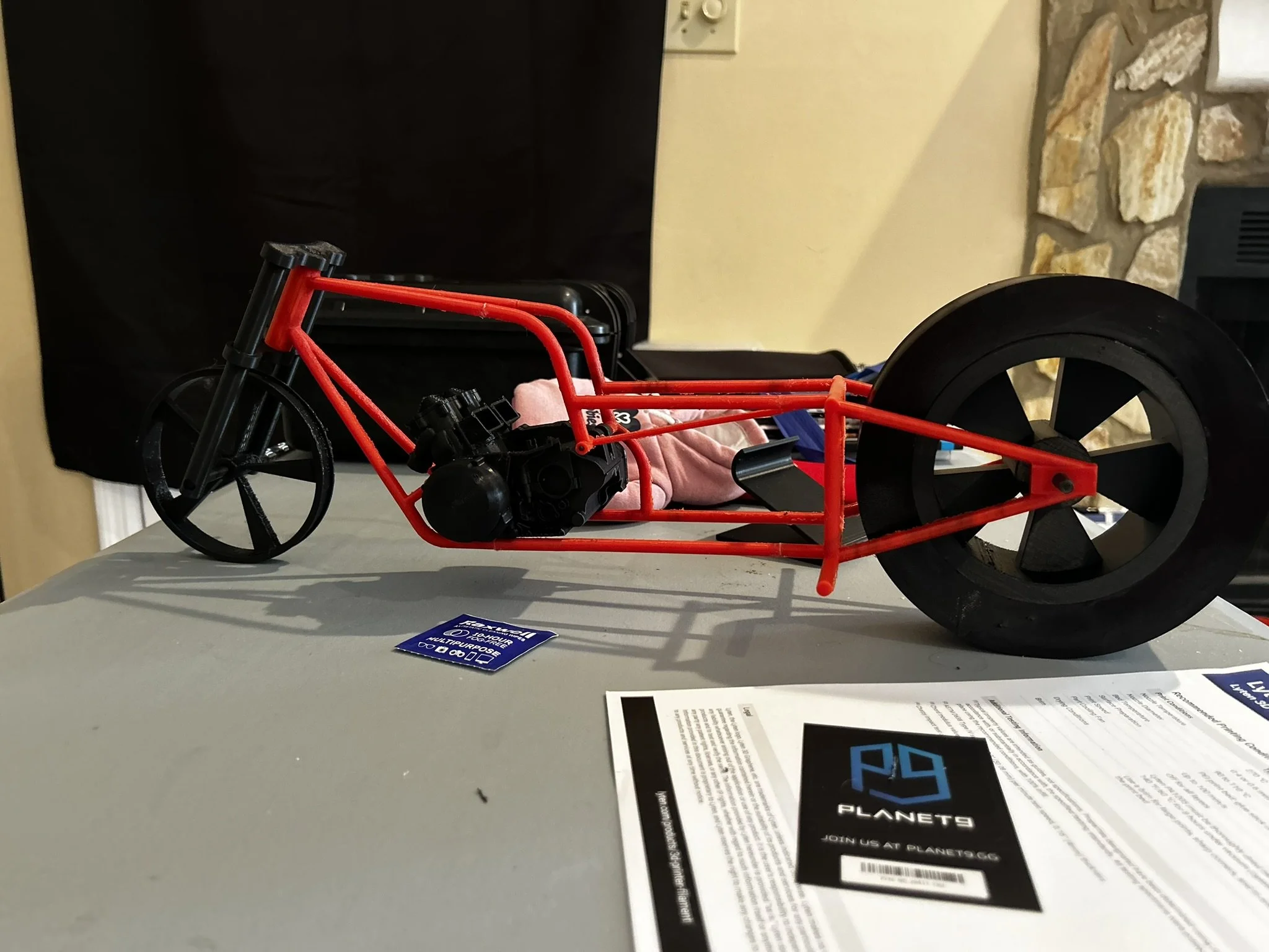A red plastic toy race car on a light-colored table, with a black background and some paper advertisements or flyers in the foreground.
