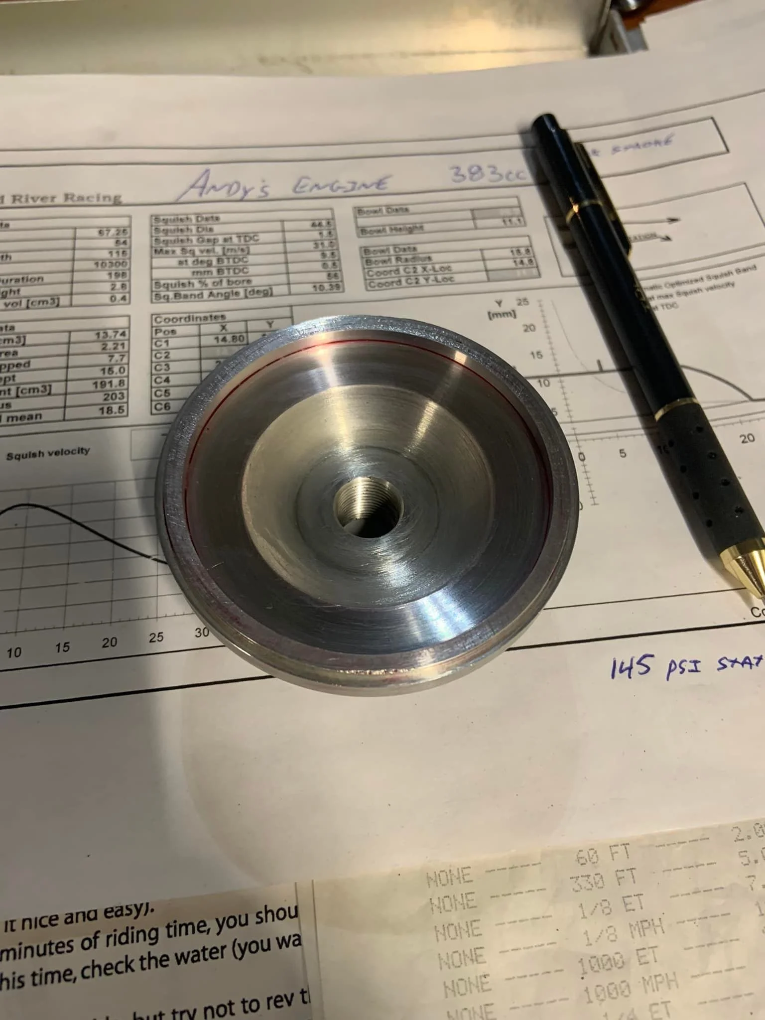 A metal motor pulley placed on top of a racing data sheet, with a pen and handwritten notes indicating 145 PSI stat pressure. The data sheet contains specifications and data for Andy's Engine 383cc, and the background shows part of a printed document or form.