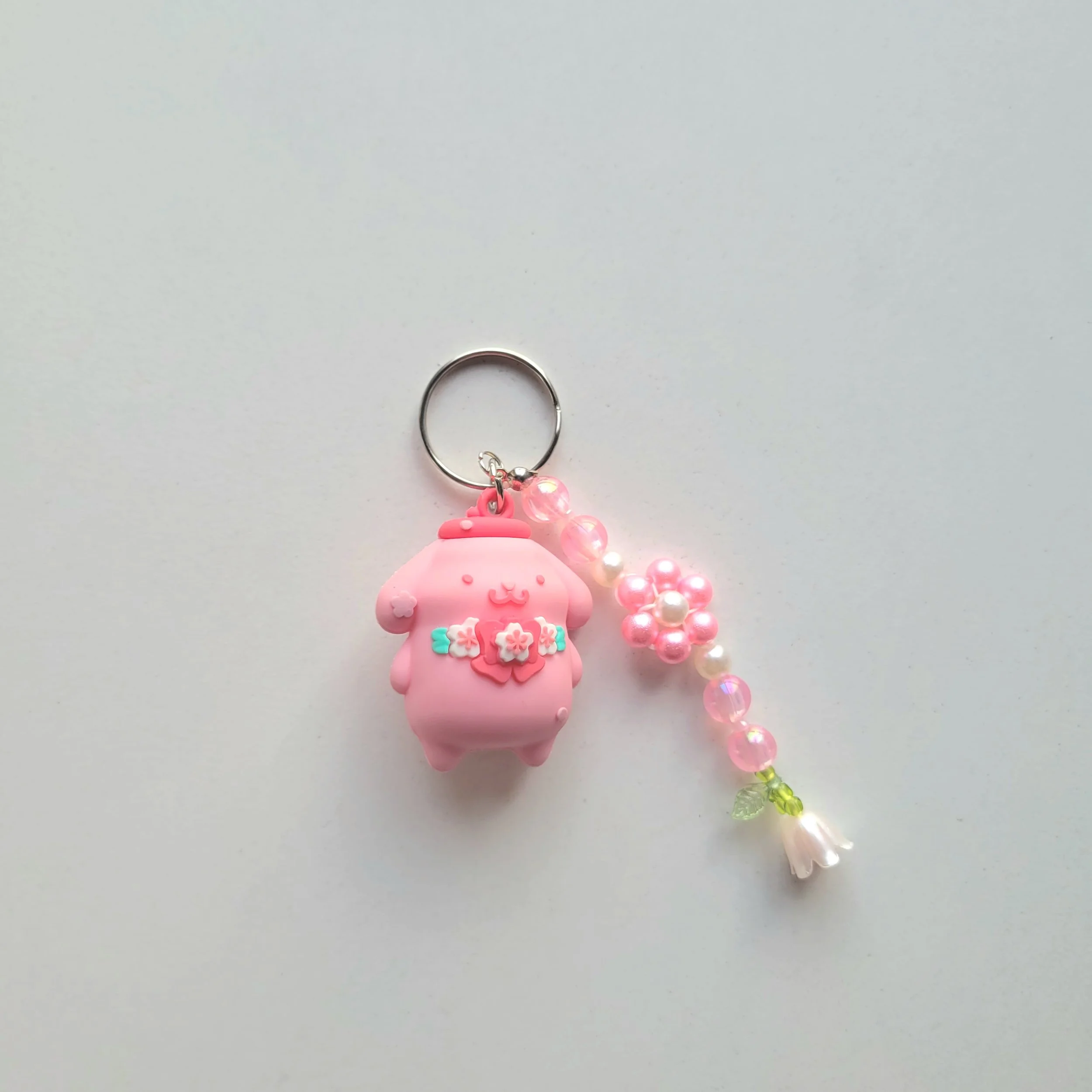 A pink baby figure keychain with a smiling face, small hands and feet, and decorated with white, pink, and aqua floral patterns, hanging from a silver keyring attached to a pink and pearl beaded chain that illustrates a flower