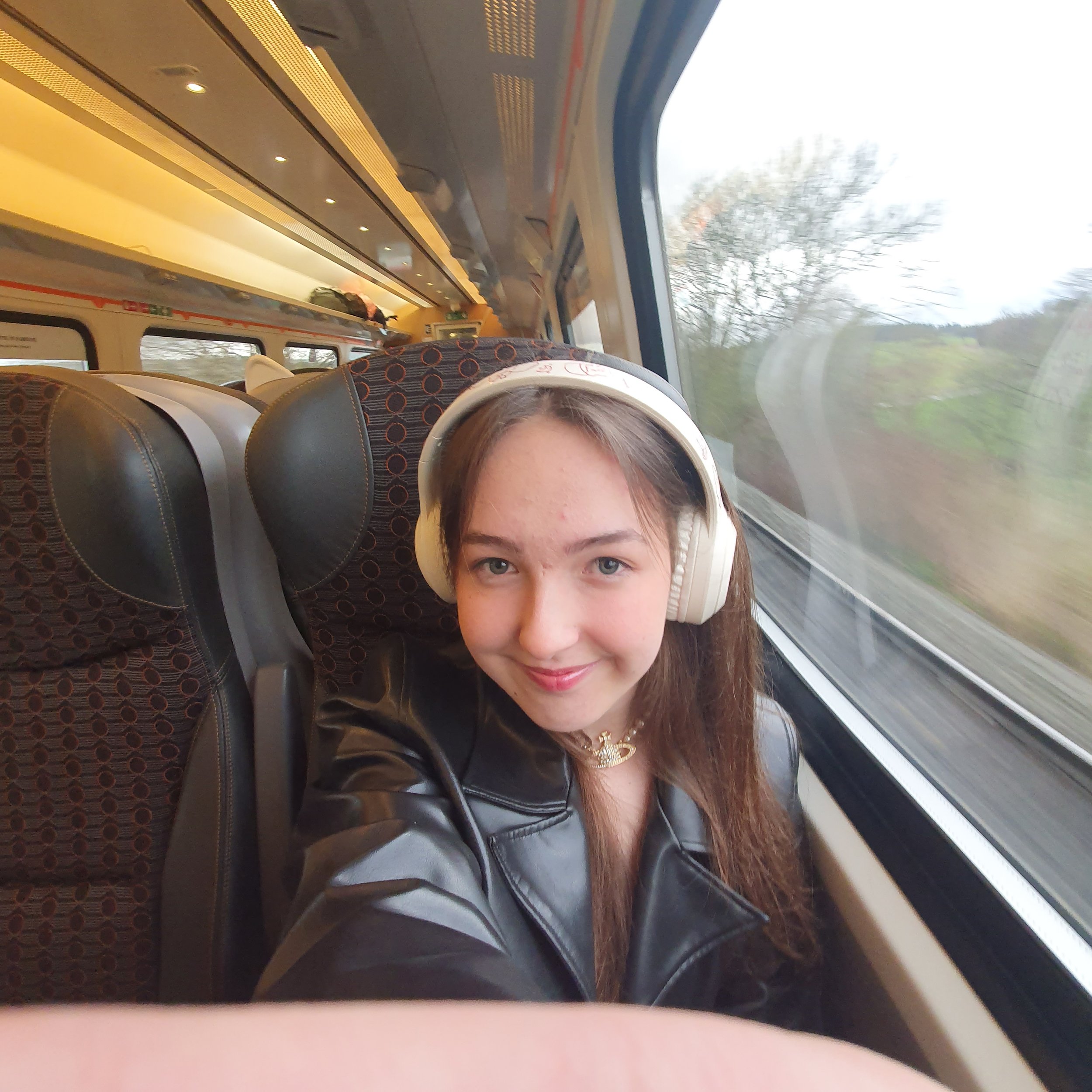 The image shows a person sitting inside a train, with a view of their face and upper body. They are wearing headphones and have long brown hair. The train interior has patterned seats, overhead luggage storage with bags, and a window showing a blurred landscape of trees and grass outside.
