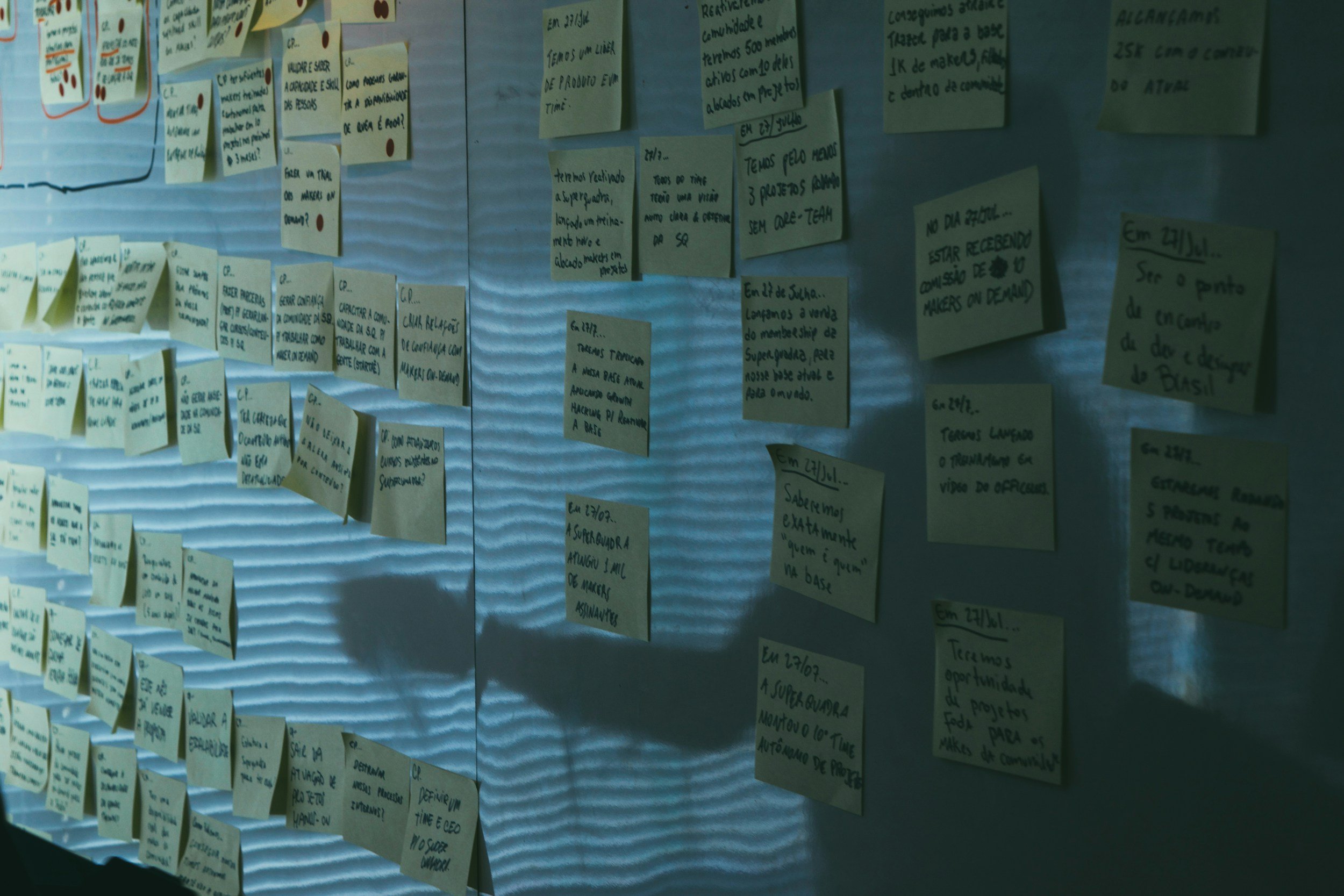 A wall with numerous yellow sticky notes with handwritten notes attached to a corrugated metal surface.
