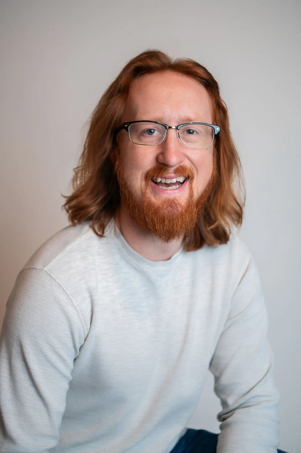 A young man with shoulder-length red hair, a beard, glasses, and a white shirt, smiling at the camera against a plain background.