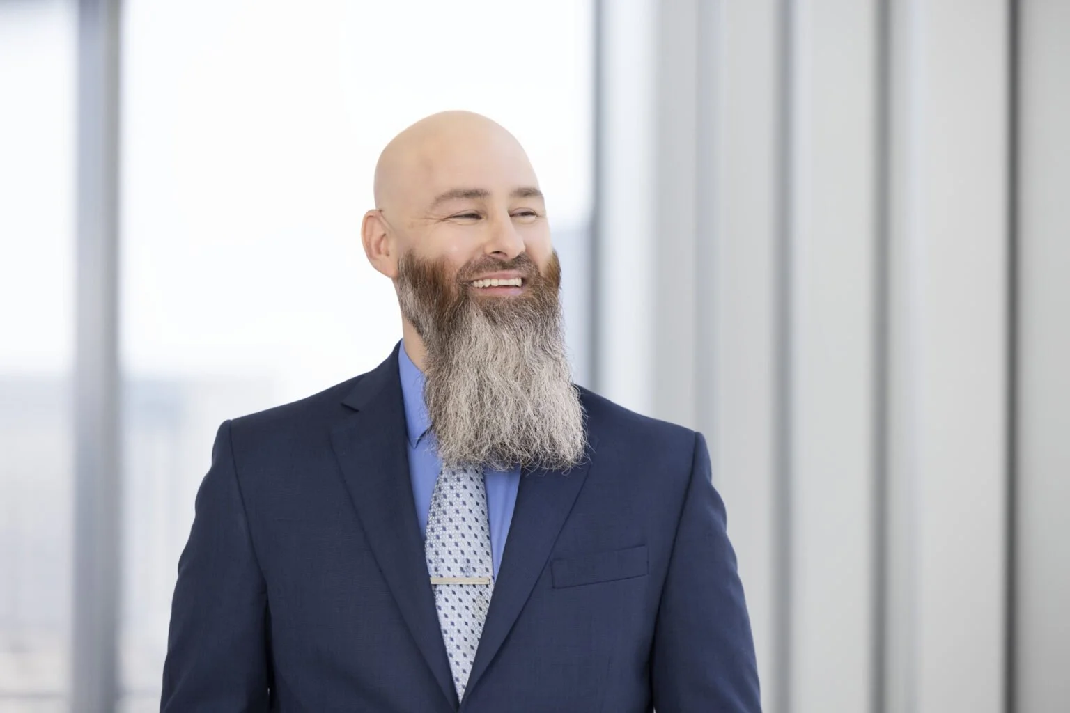 A smiling bald man with a gray beard in a navy suit, light blue shirt, and patterned tie standing in a modern office with large windows.
