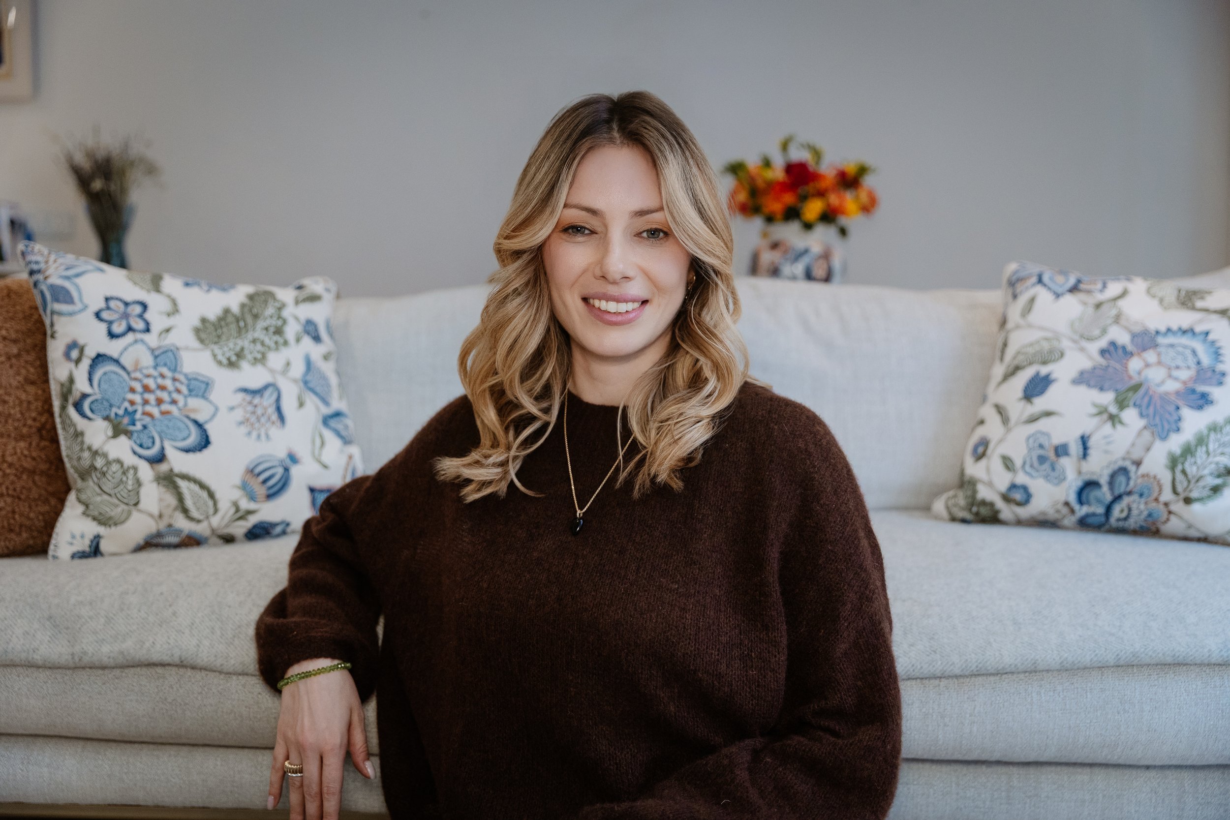 A woman with long wavy blonde hair sitting on a sofa, smiling at the camera. She is wearing a dark brown sweater, a necklace, and a bracelet. Behind her, there are decorative pillows with floral patterns and a vase with flowers on a table.