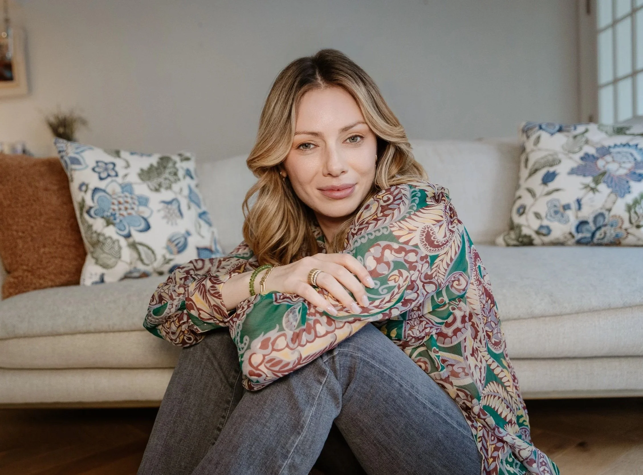 A woman with blonde hair and a patterned blouse sitting on a beige couch with floral cushions, smiling softly.