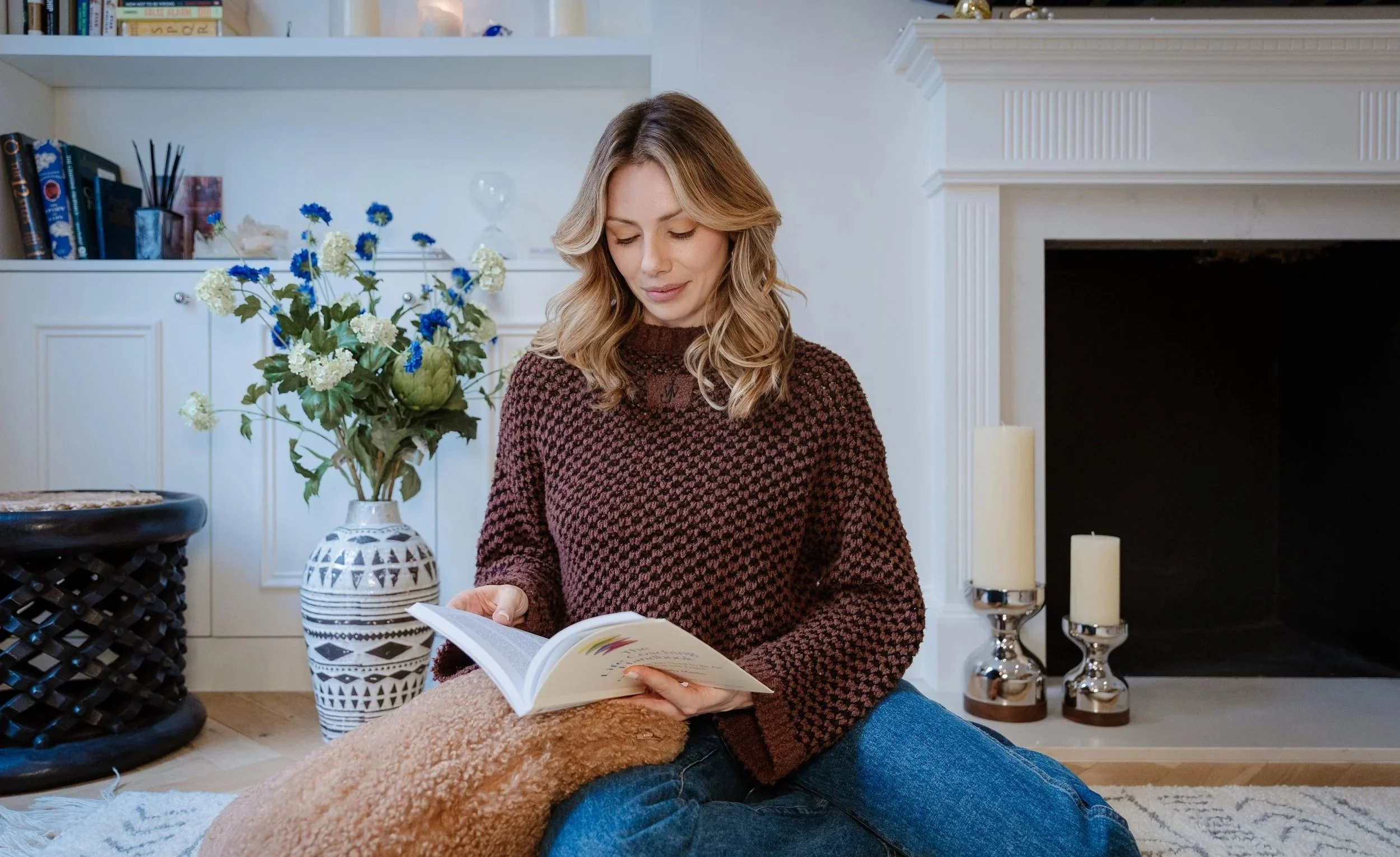 A woman with wavy blonde hair sits on the floor, reading a book. She wears a brown patterned sweater. Behind her, there is a white fireplace with candles on the mantel, a large bouquet of white and blue flowers in a decorative vase, and a white bookshelf with various books and decorations. The room has a cozy, modern decor with white walls and a light-colored rug.