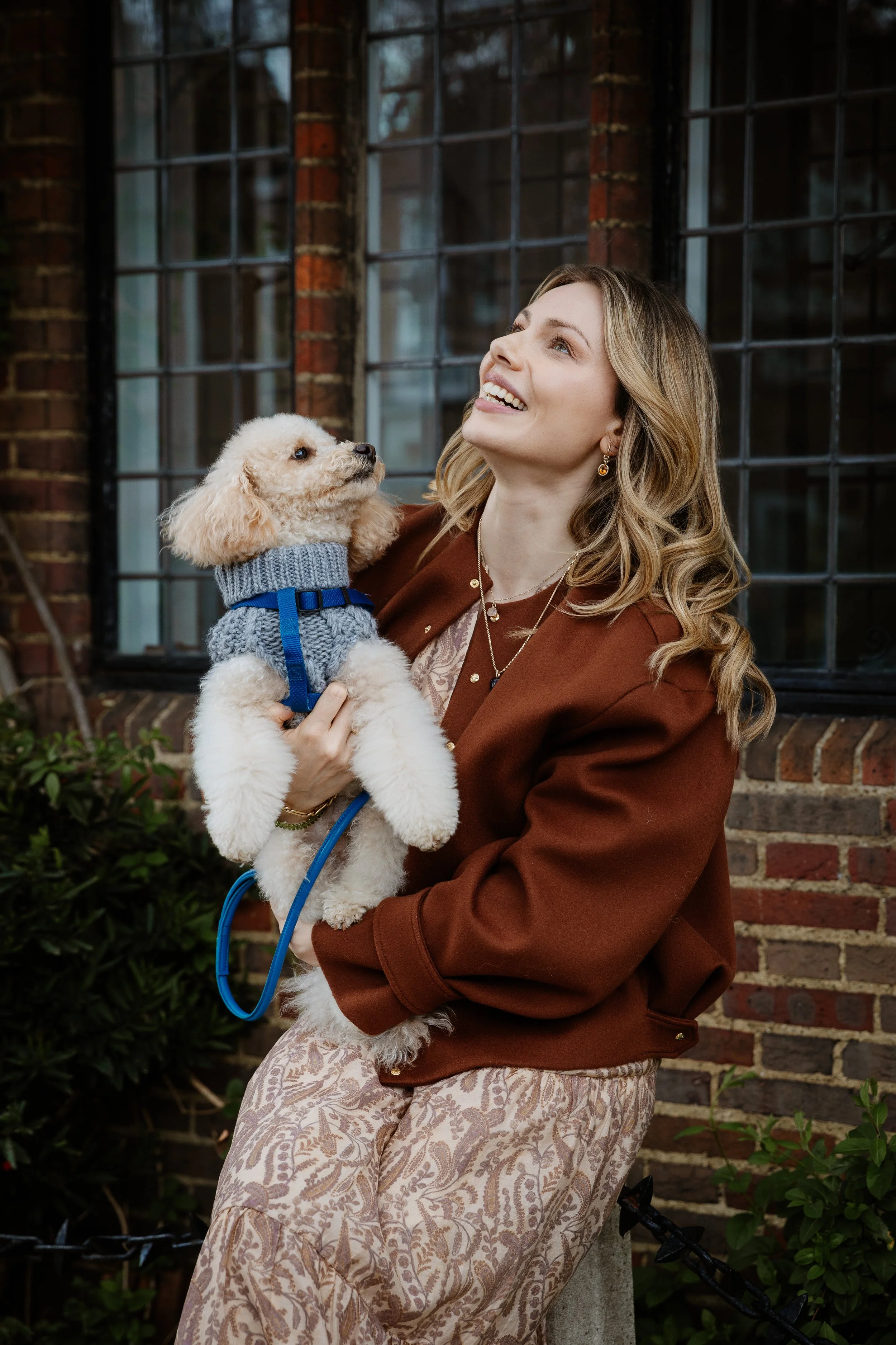 Woman smiling and looking up while holding a small curly-haired dog wearing a gray sweater and harness, sitting outdoors in front of a brick building with large windows.