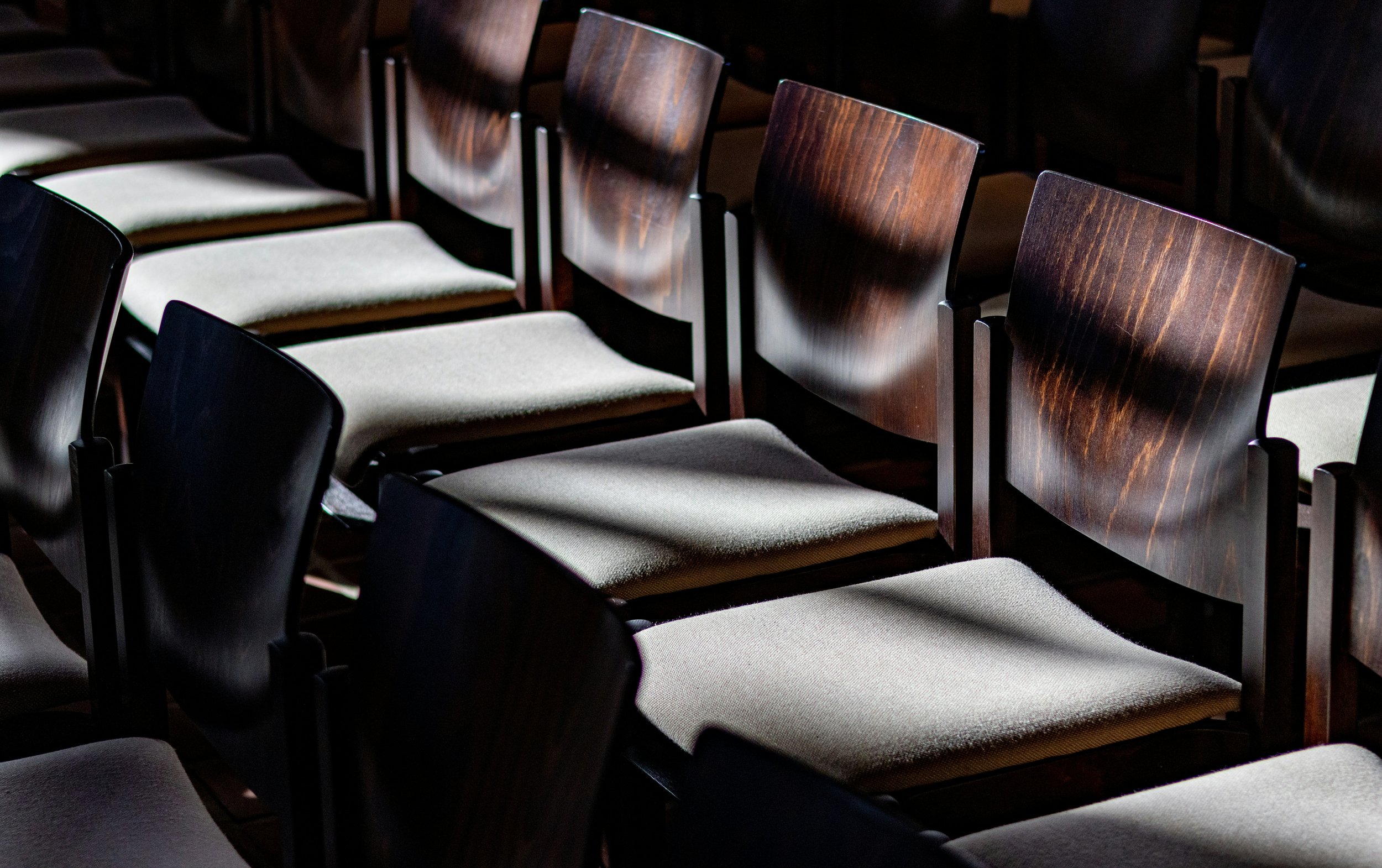 Rows of wooden chairs with padded seats in a dimly lit room, possibly a theater or auditorium.