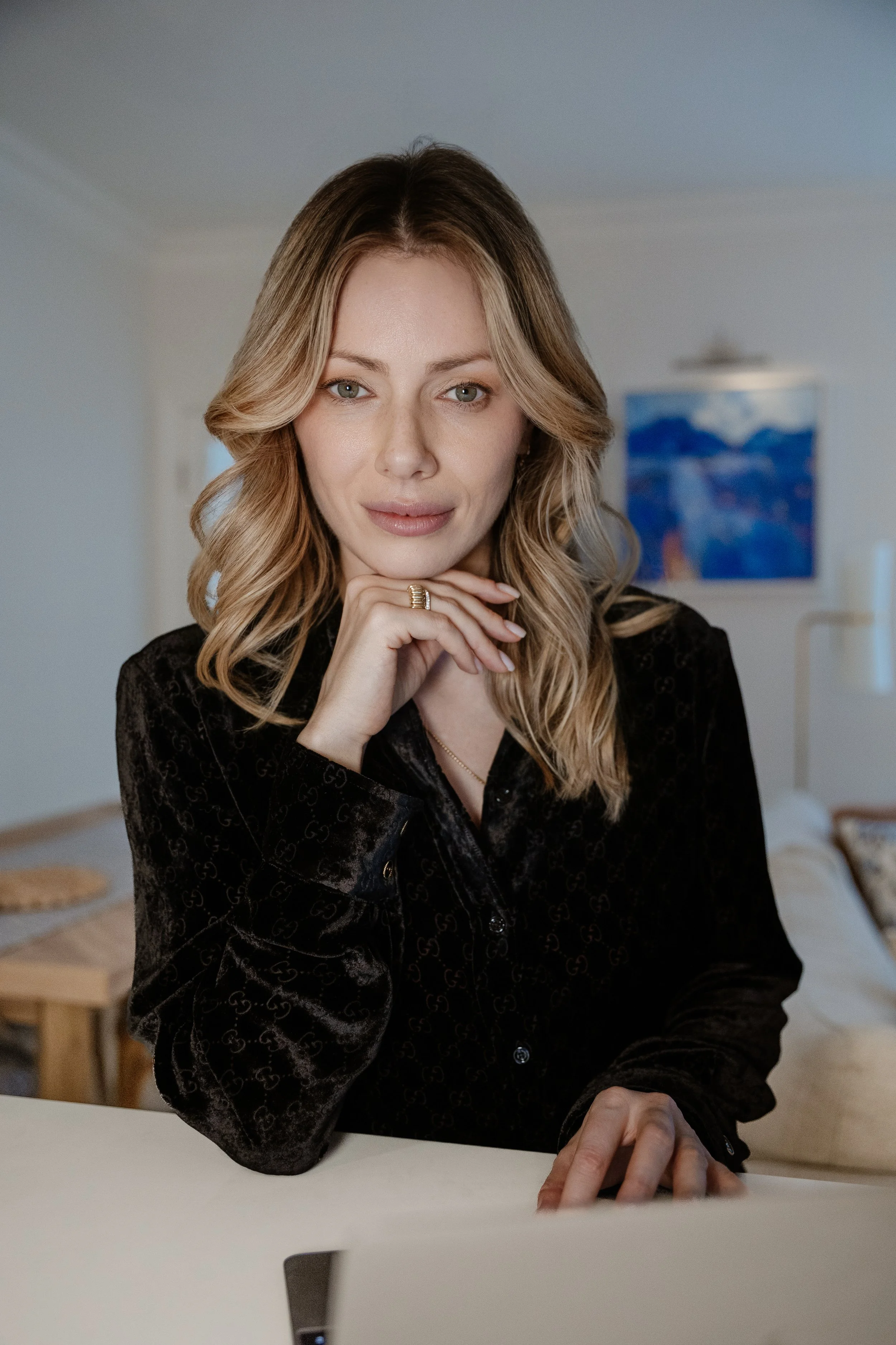 A woman with blonde hair and light skin is smiling softly while resting her chin on her hand. She wears a black, long-sleeved, patterned top and a gold ring on her finger. The background includes a colorful landscape painting and some furniture.