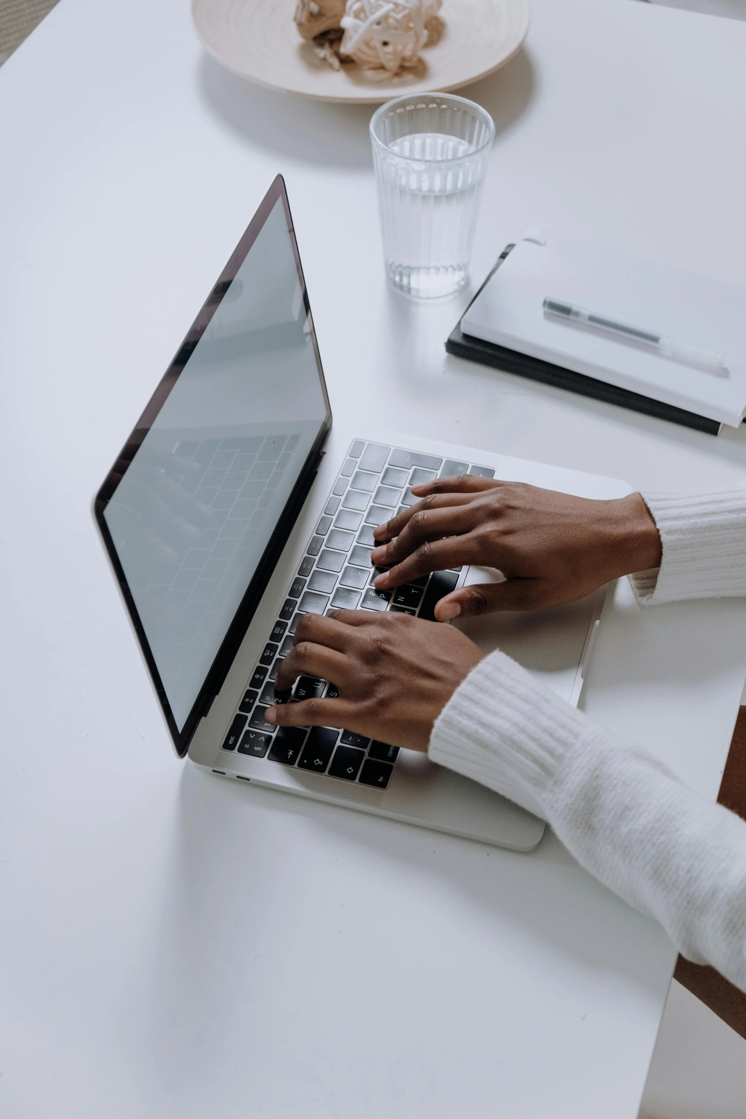 A person wearing a white sweater typing on a laptop keyboard at a white table with a glass of water, a notepad, a pen, and a plate of food.