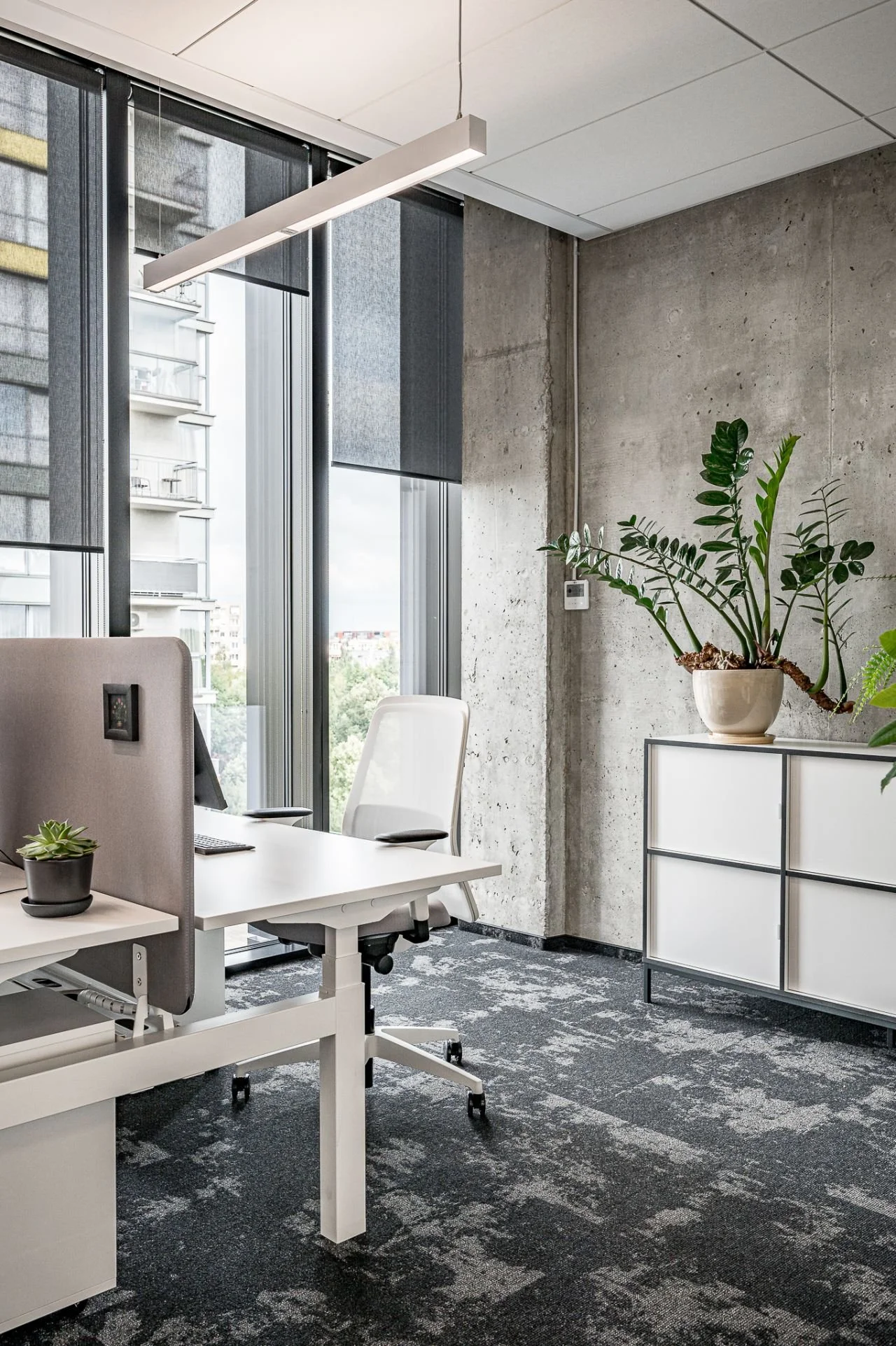 Modern office space with a white desk, computer, and an ergonomic chair, beside large windows with black blinds and a concrete wall with a potted plant on a white cabinet.